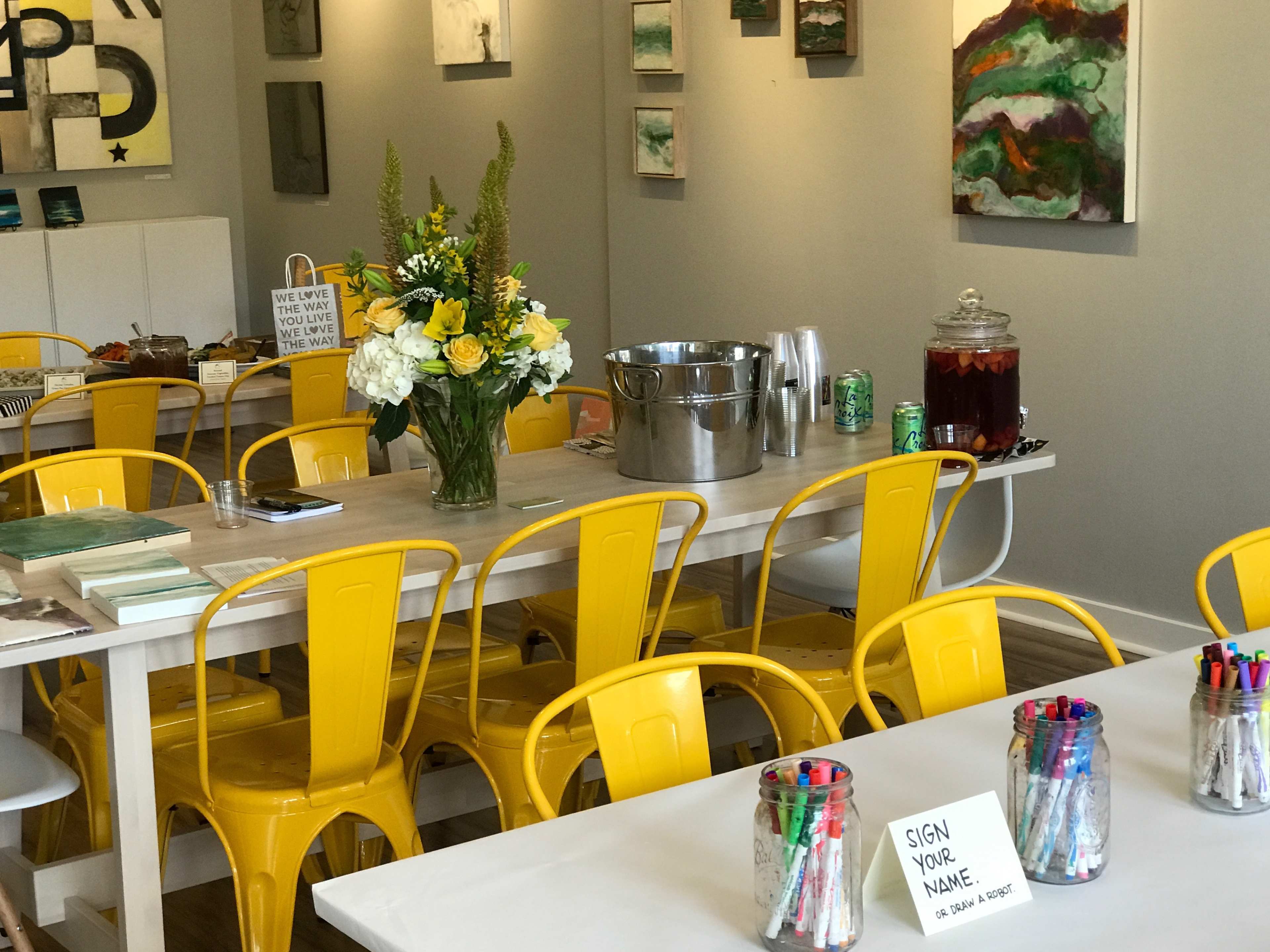 A modern, brightly lit room features yellow metal chairs around a long wooden table, adorned with a flower vase and various beverages, alongside a sign inviting guests to sign their names.