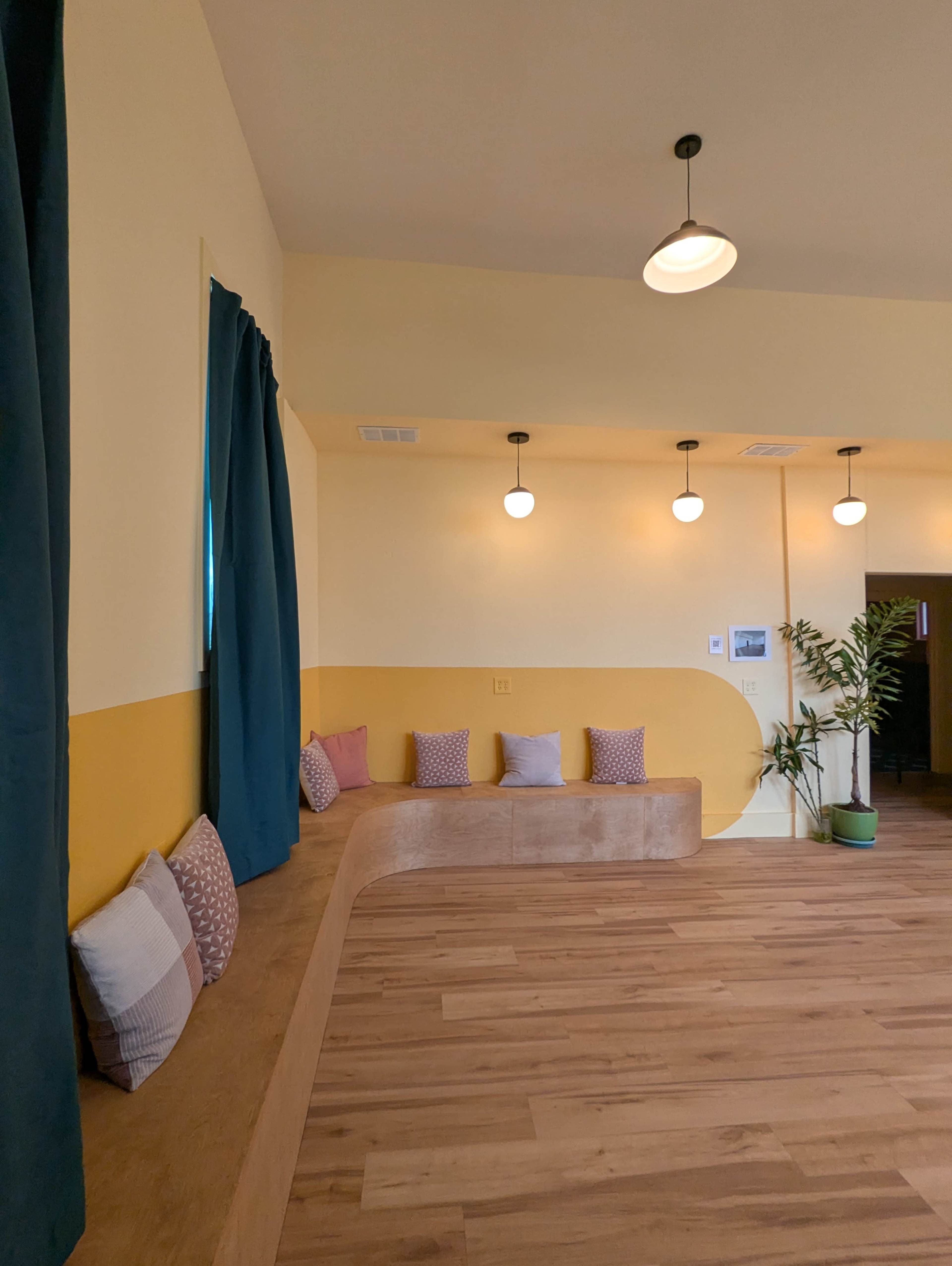 The image shows a cozy interior space with a wooden bench lined with cushions against a wall featuring yellow and pale yellow paint, alongside large windows covered with blue curtains and hanging light fixtures.