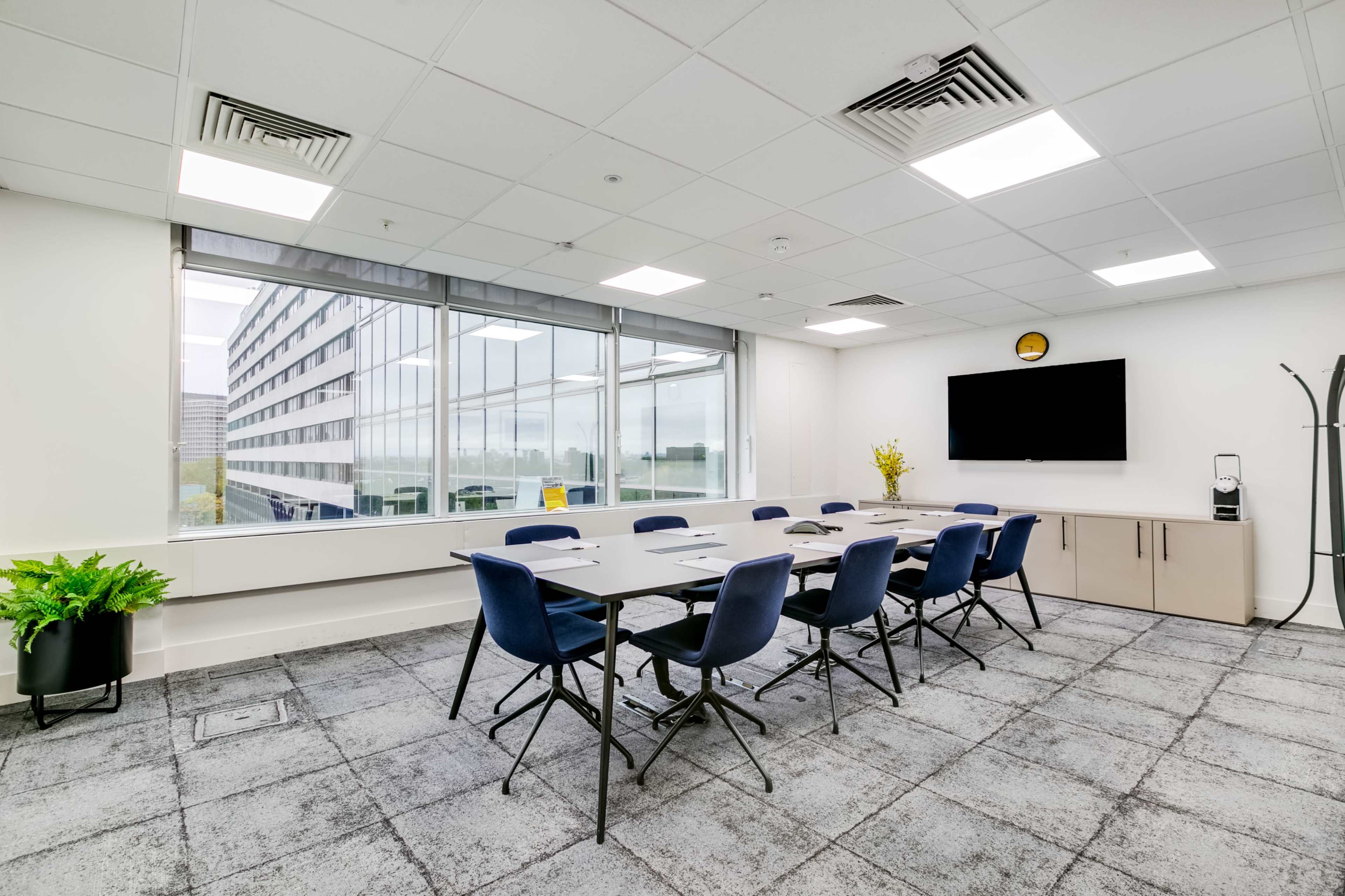 A modern conference room features a long table surrounded by chairs, a large window with a view, and a television mounted on the wall.
