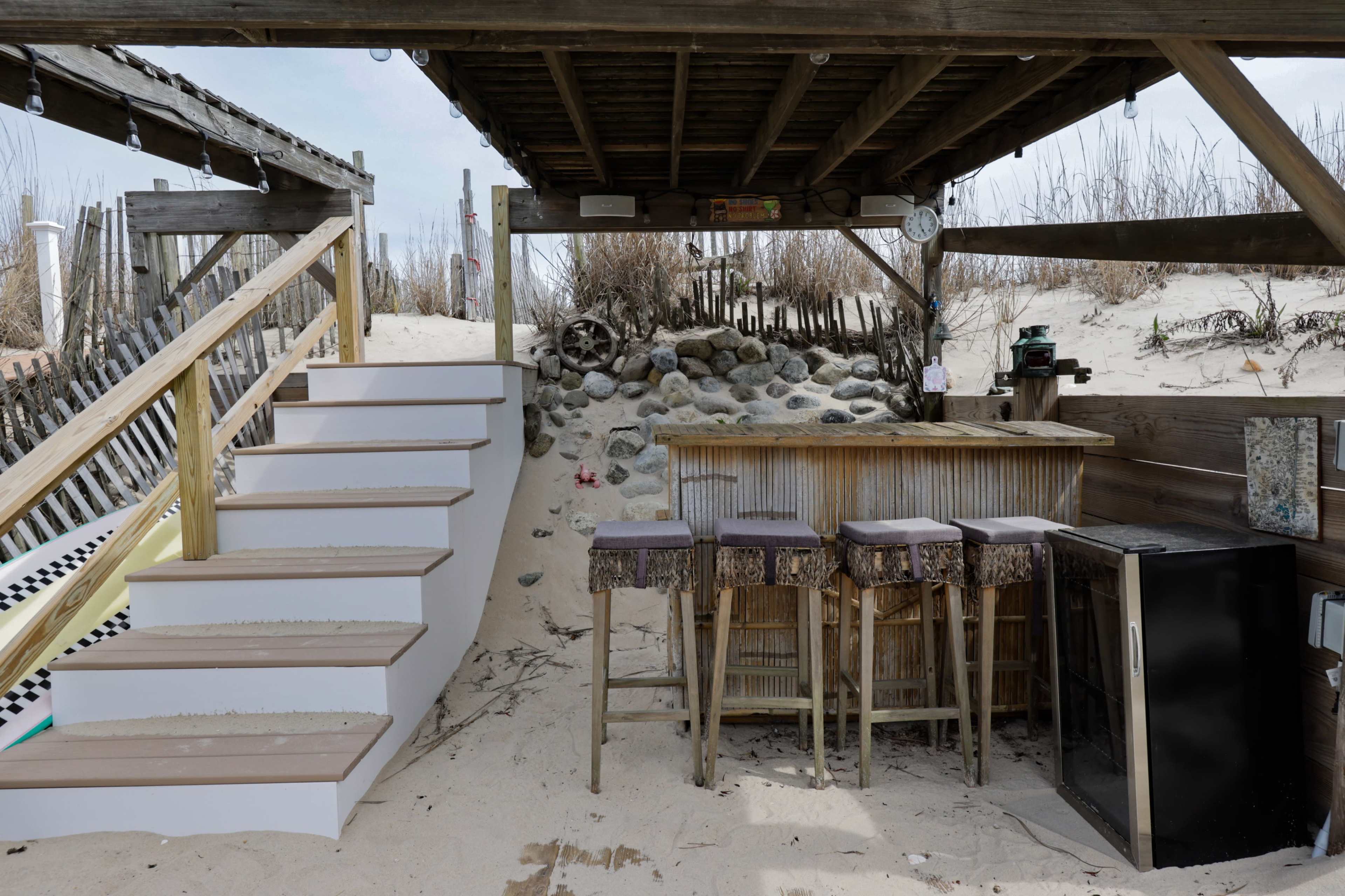 The image shows an outdoor bar area with wooden stools and a refrigerator, situated under a rustic shelter near sandy steps leading up to a deck.