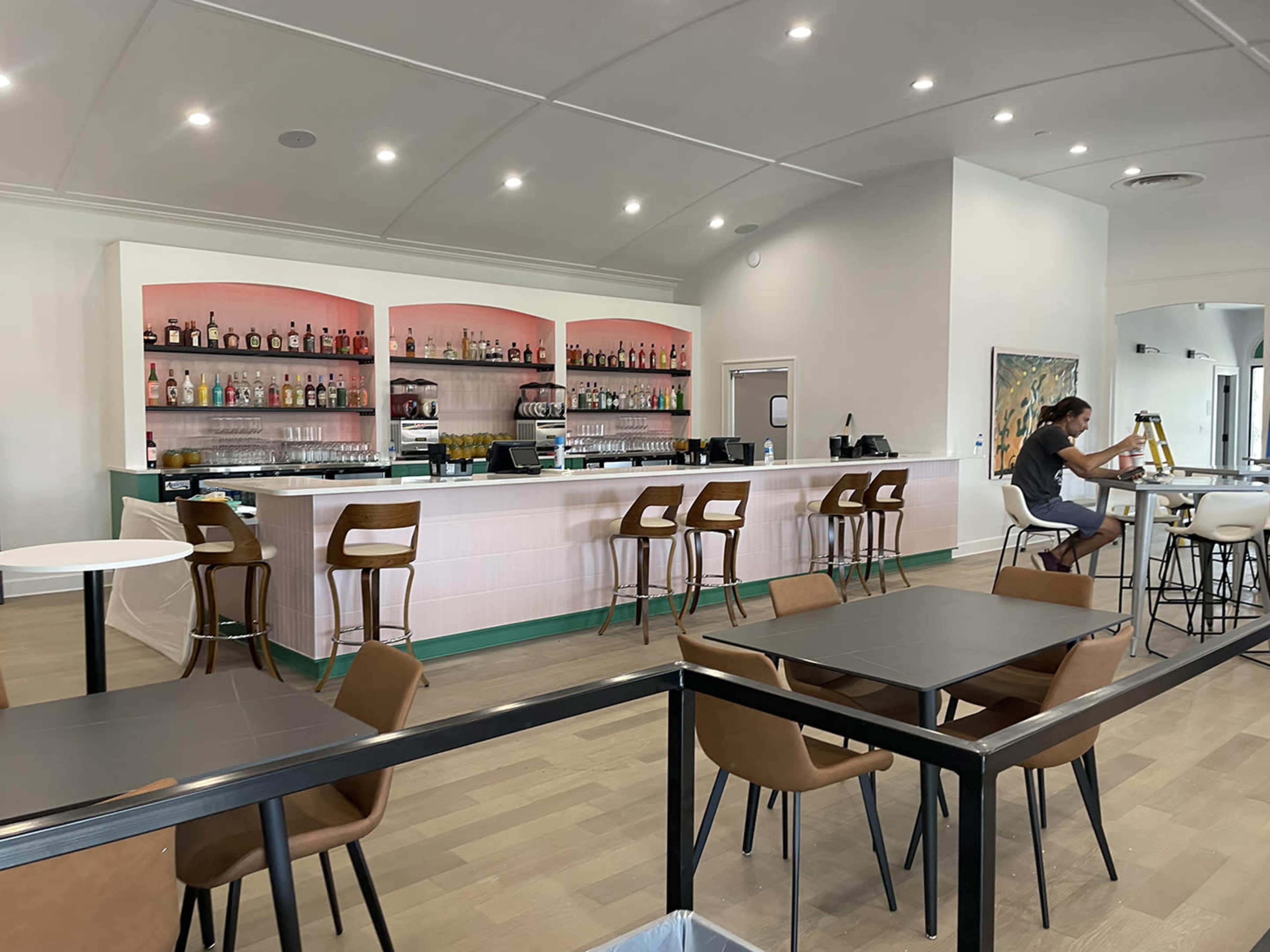 The image shows a modern bar area with a counter stocked with various bottles and several seating tables and chairs in a well-lit space.