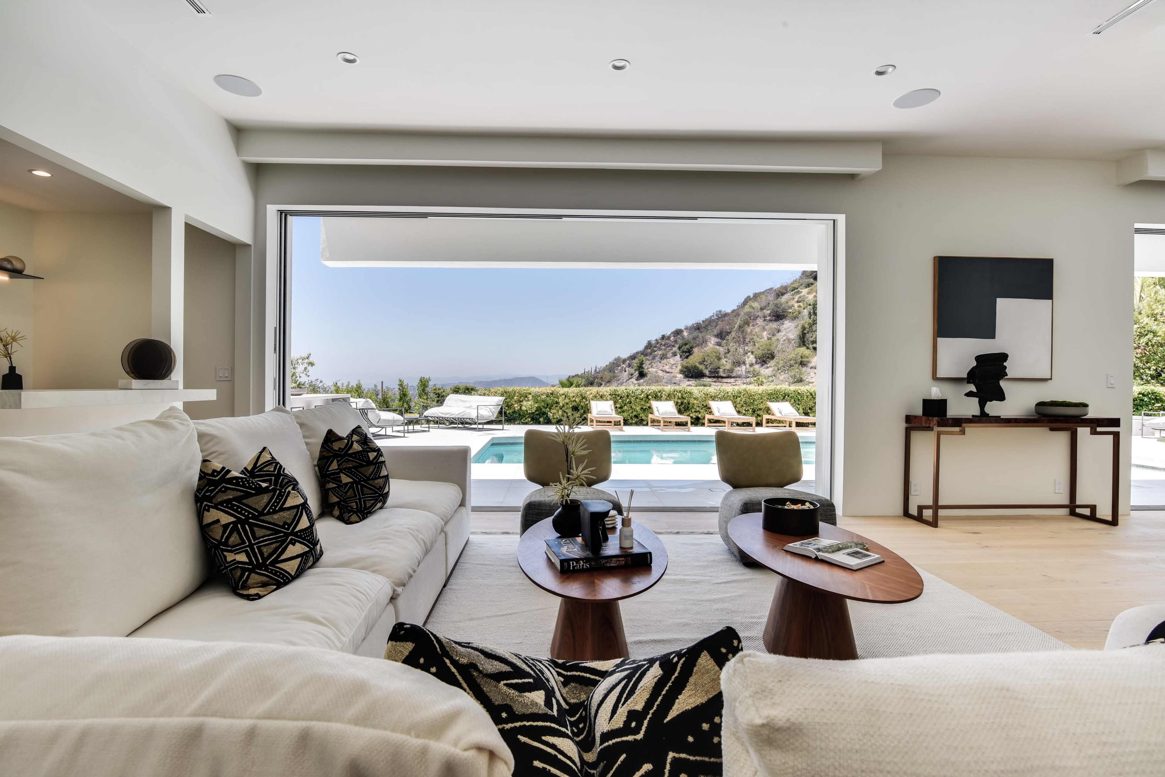 A modern living room features a white sofa facing a large glass window that leads to a pool area surrounded by outdoor furniture and scenic hills.