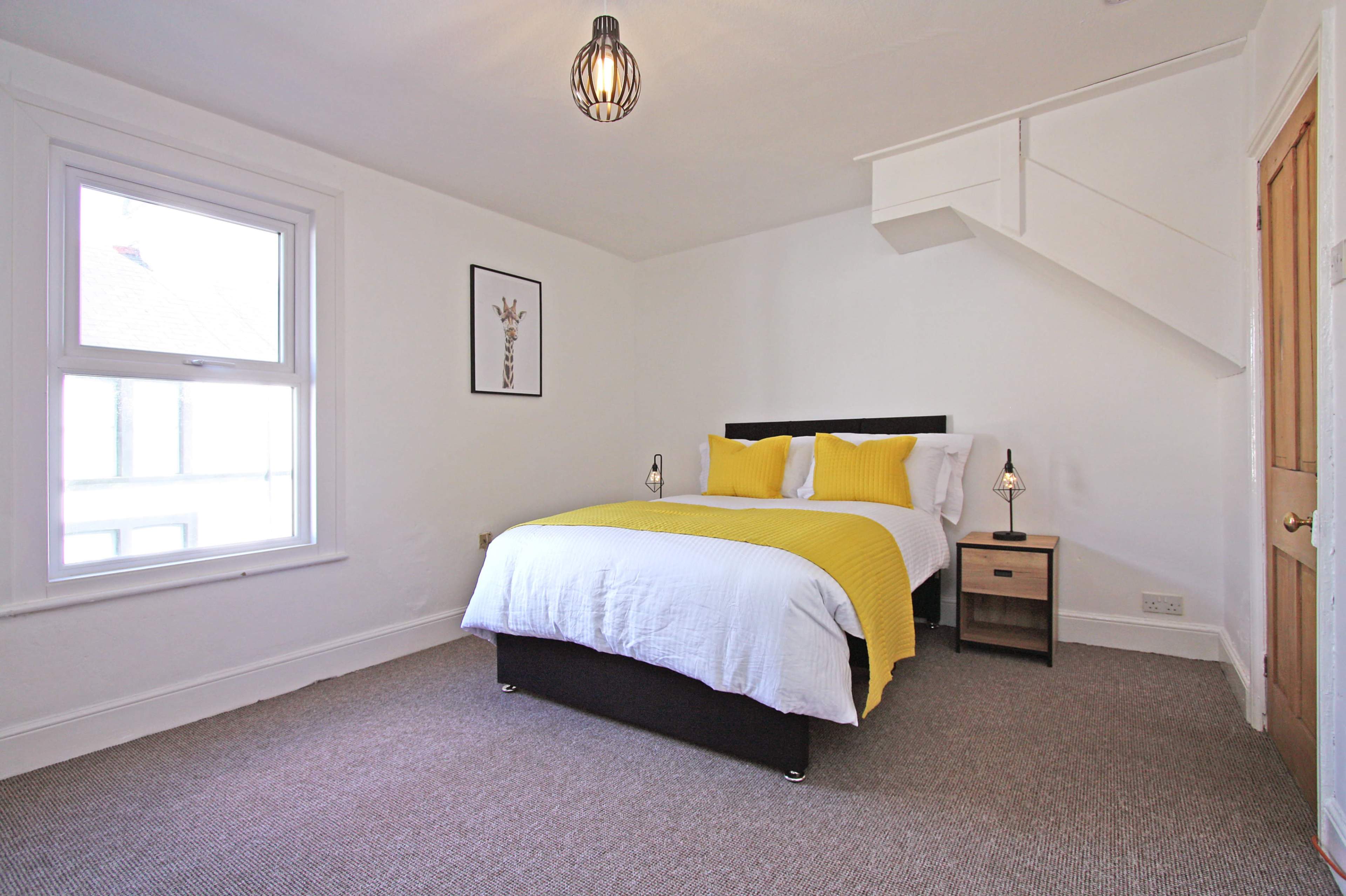 The image shows a bright bedroom with a double bed dressed in white linens and yellow accents, a nightstand with lamps, and a window providing natural light.