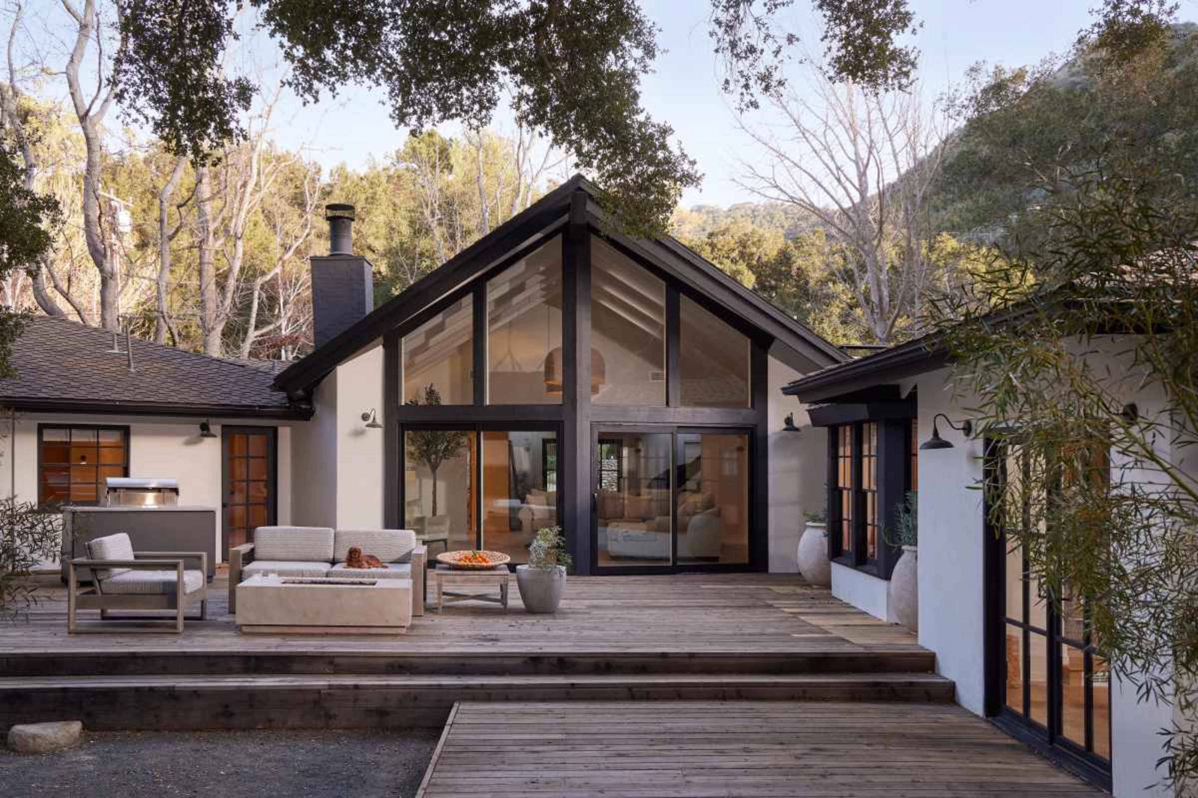 A modern house features large glass windows, wooden decks, and a natural setting with trees in the background.