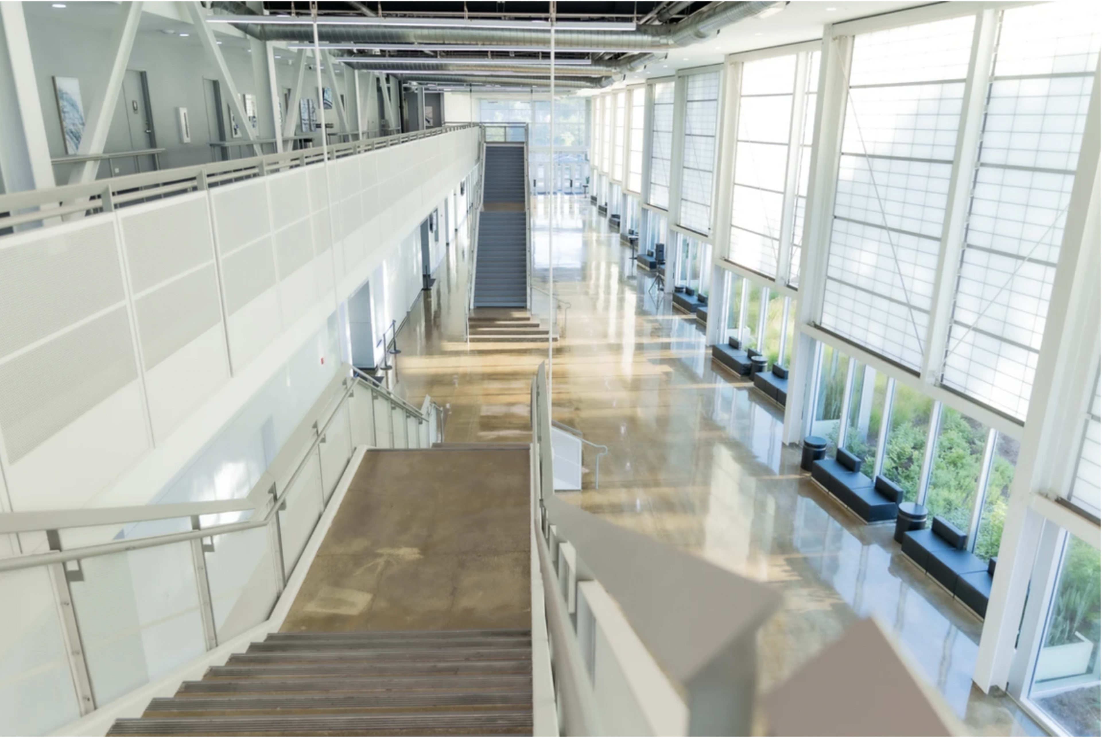 A spacious interior of a modern building with large windows, stairs leading down to a lower level, and potted plants along the corridor.