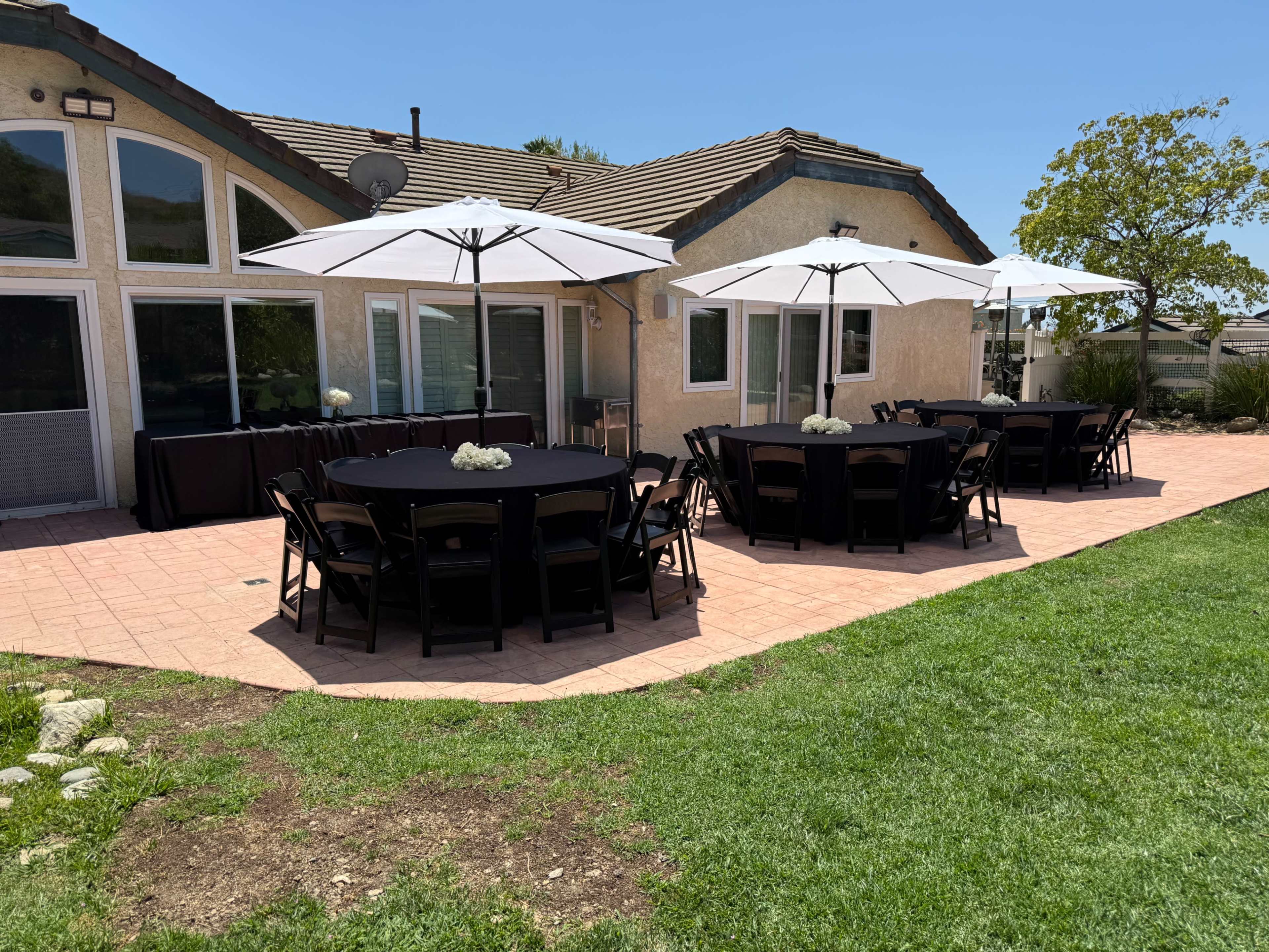 A patio area features round tables with black tablecloths and white umbrellas, surrounded by a well-manicured lawn and a house in the background.