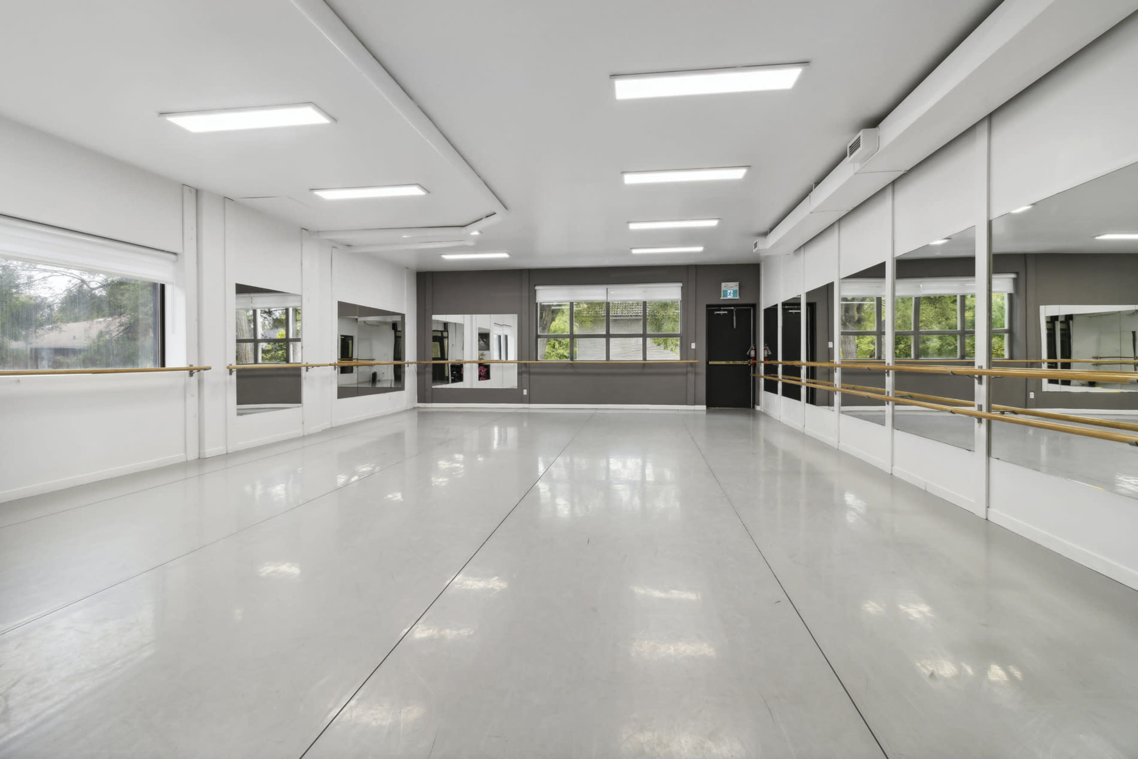 A spacious dance studio with mirrored walls and a wooden ballet bar, featuring large windows that let in natural light.