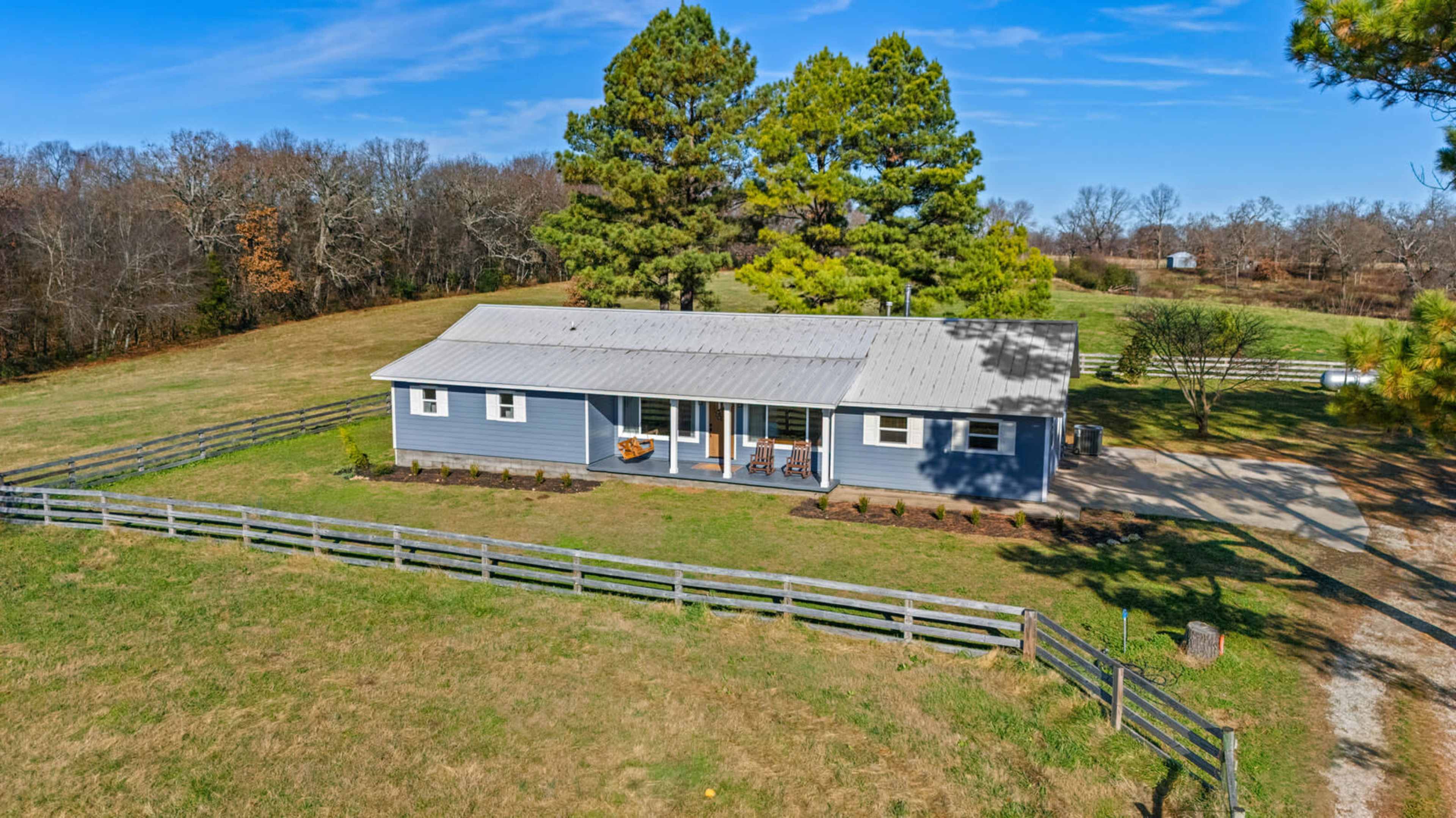 A single-story gray house with a metal roof sits on a grassy field, surrounded by a wooden fence and tall trees under a clear blue sky.