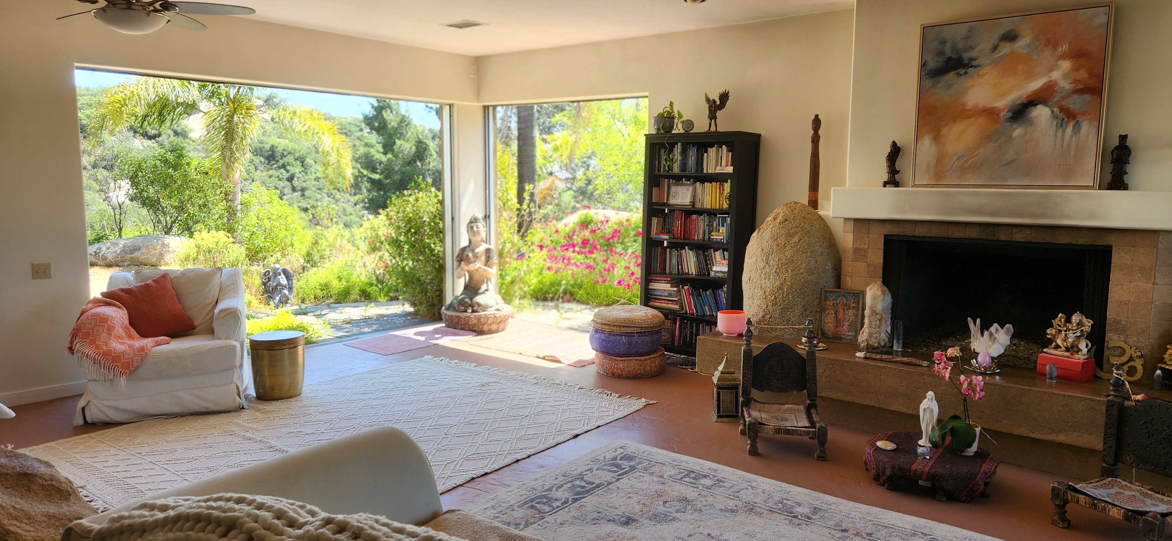A spacious living room with large sliding glass doors open to a vibrant garden, featuring comfortable seating, bookshelves, and decorative elements.