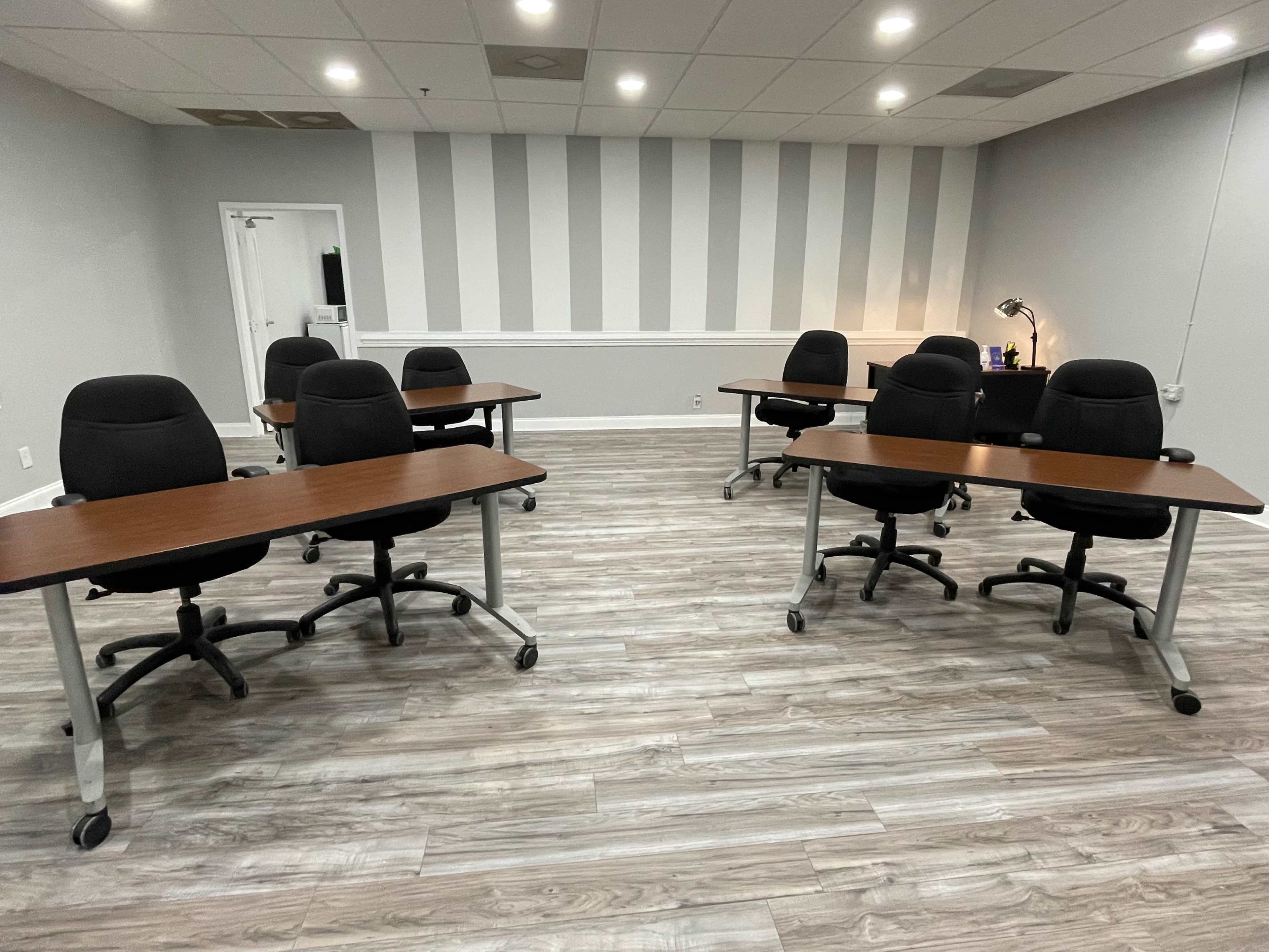 A spacious meeting room with several black office chairs arranged around wooden tables on a light-colored wooden floor.