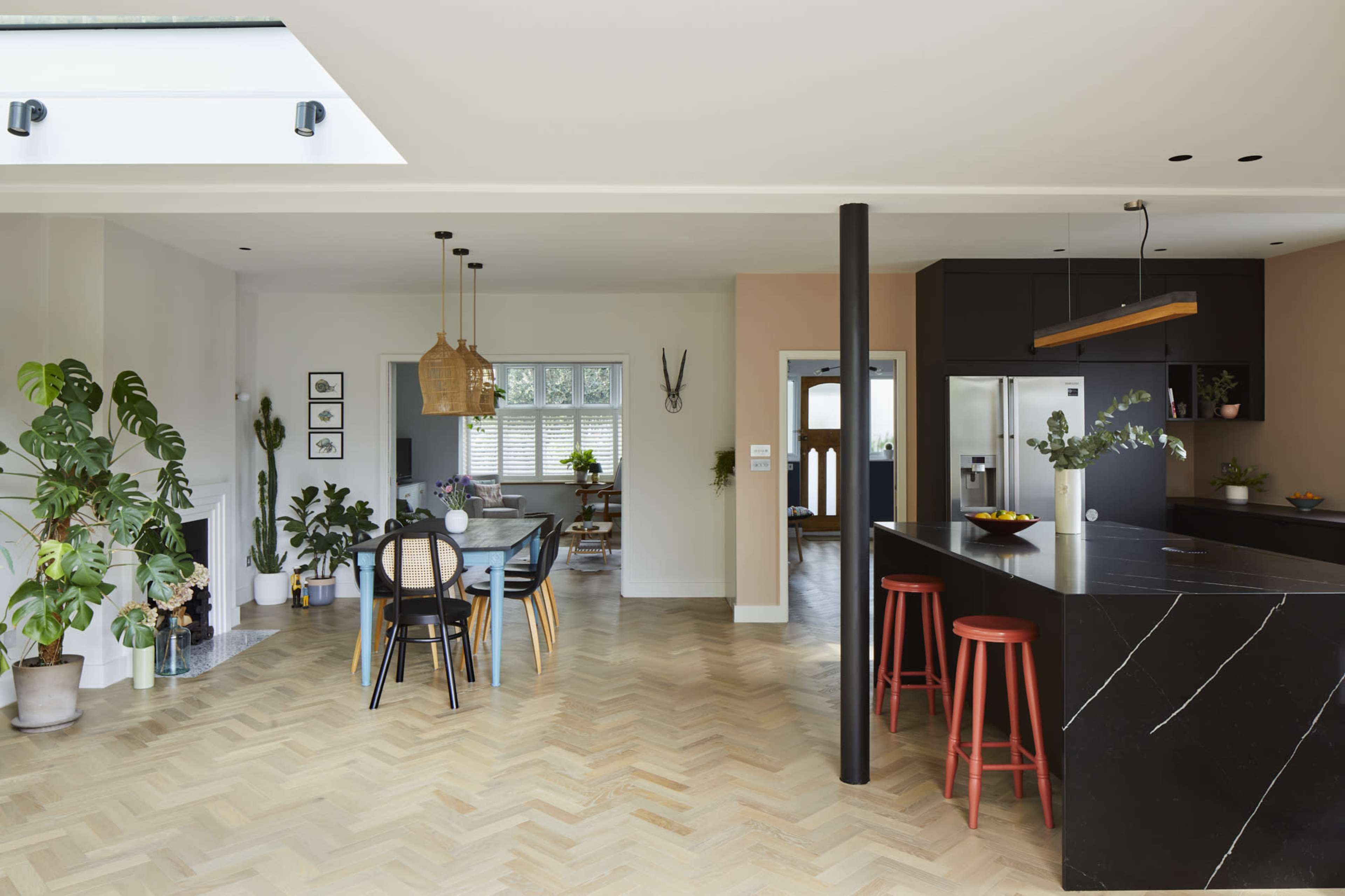 A modern kitchen and dining area feature a large island, black cabinetry, a wooden dining table, and various indoor plants.