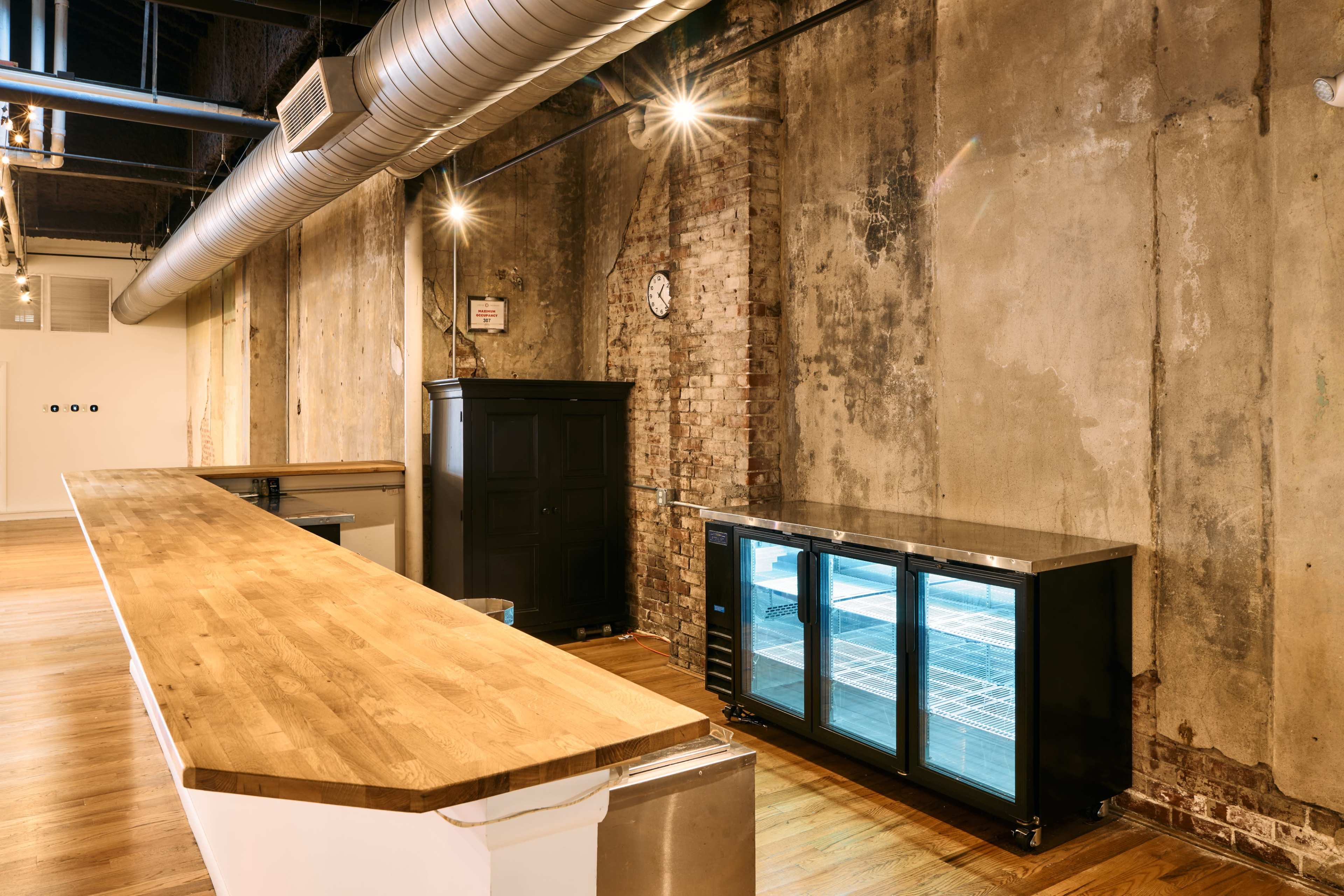 The image shows a modern industrial space with exposed brick and concrete walls, featuring a long wooden counter and a glass-fronted refrigerator.