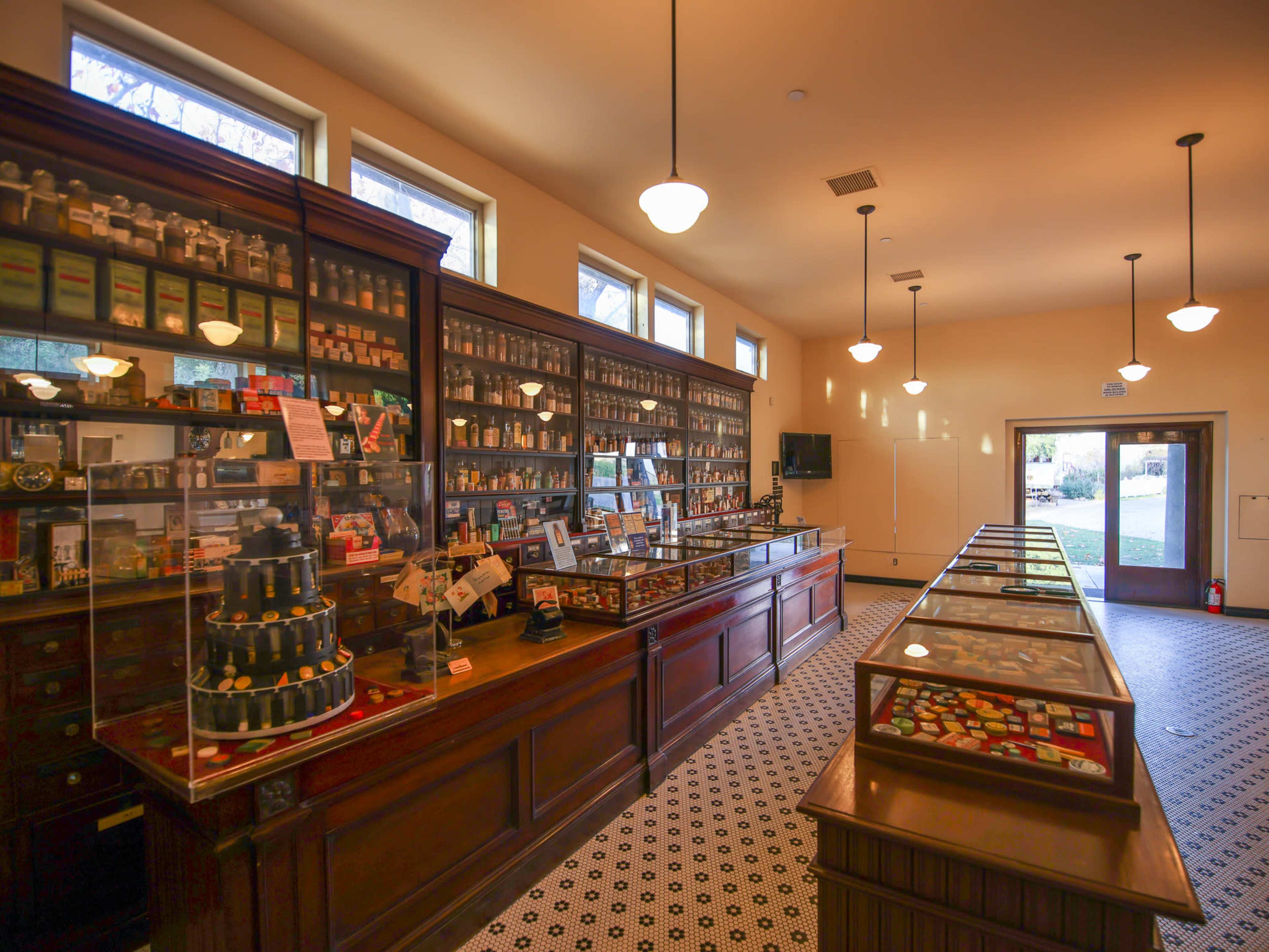 A vintage apothecary shop interior with wooden displays, glass cases filled with jars and various items, and large windows along one wall.