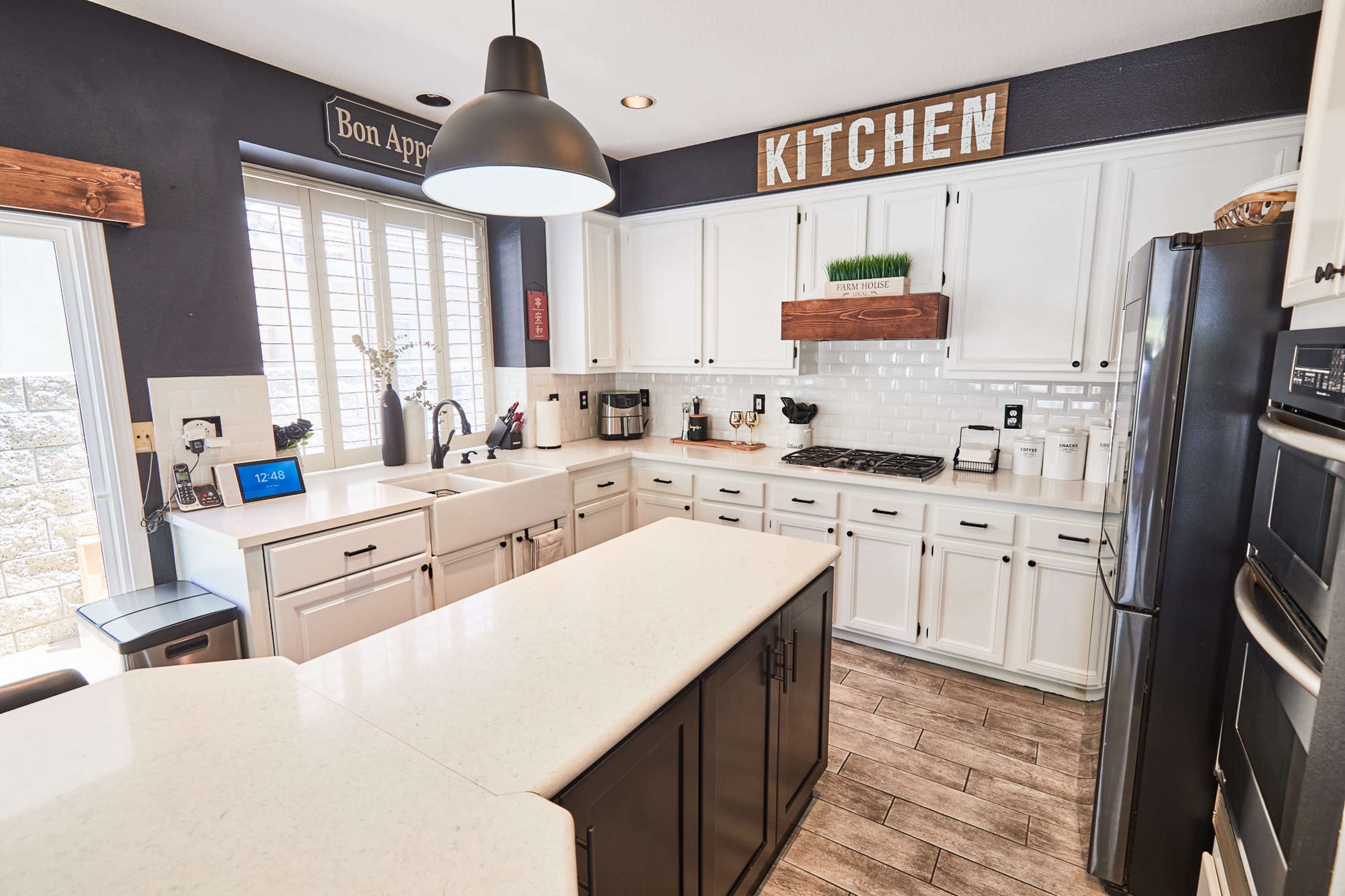 A modern kitchen features white cabinets, a central island, and large windows with natural light illuminating the space.