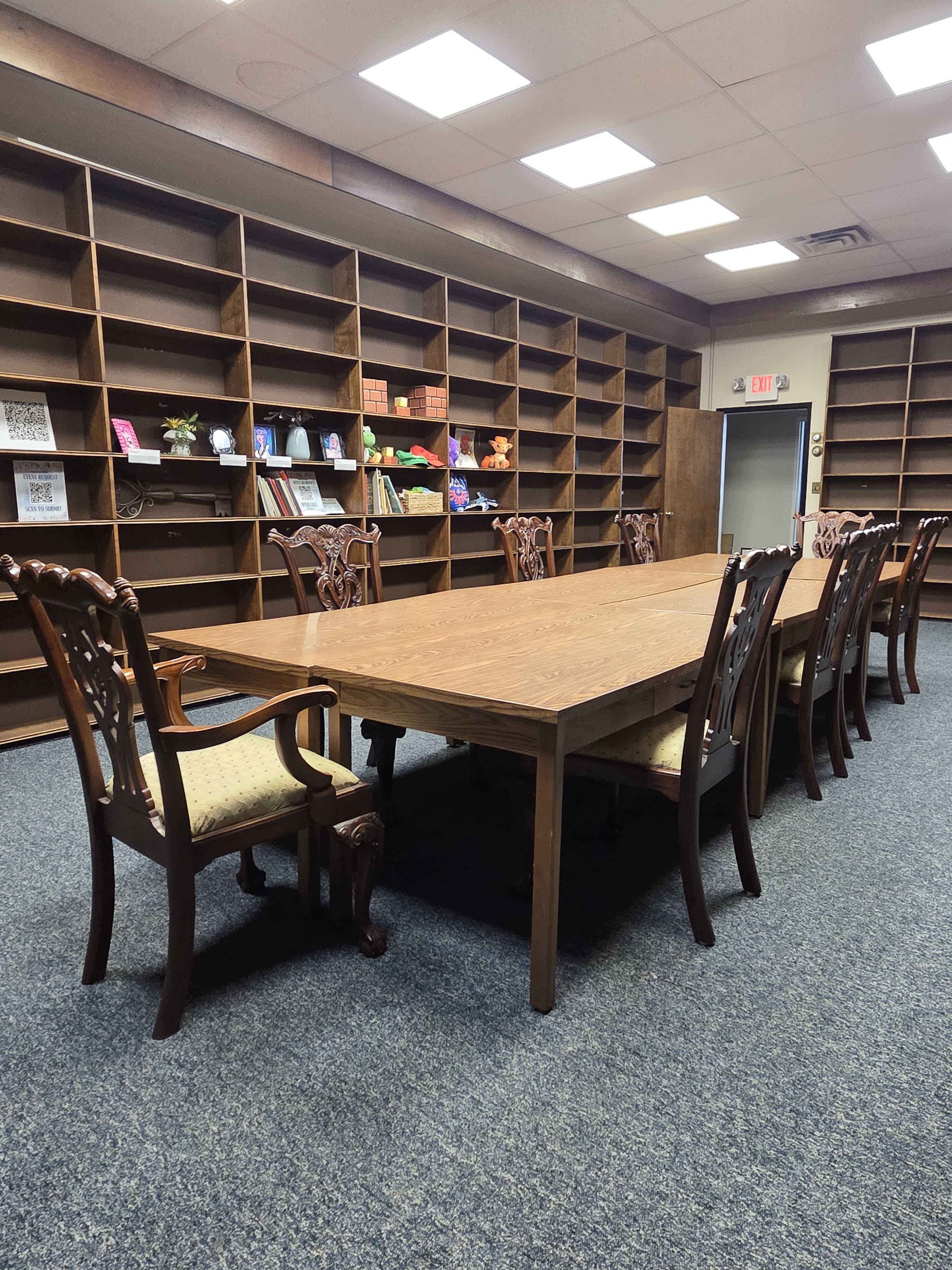 A long wooden table surrounded by carved chairs is positioned in a room with empty bookshelves lining the walls.