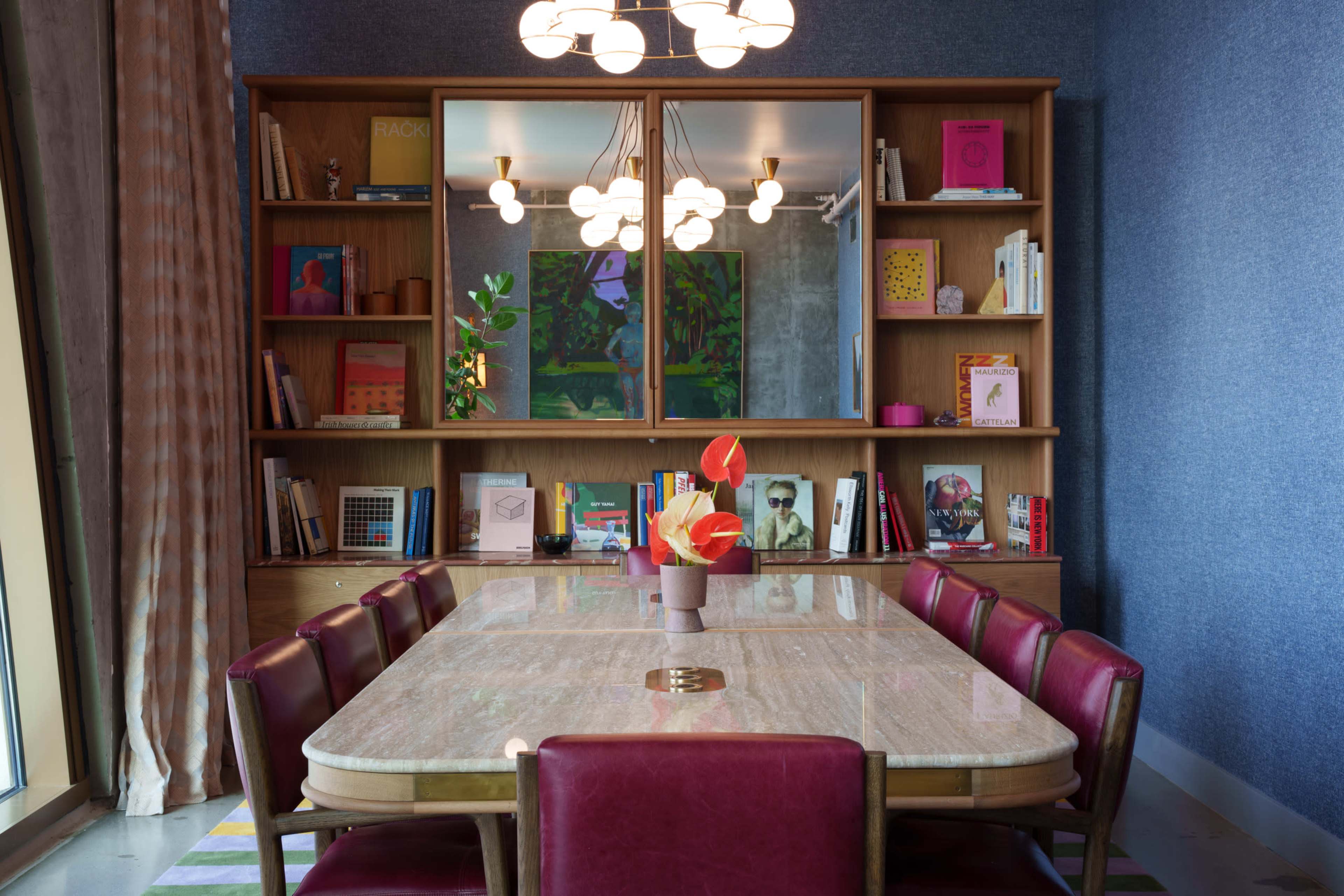 A modern dining room with a large table surrounded by burgundy chairs, a bookshelf filled with colorful books in the background, and a decorative arrangement of flowers on the table.