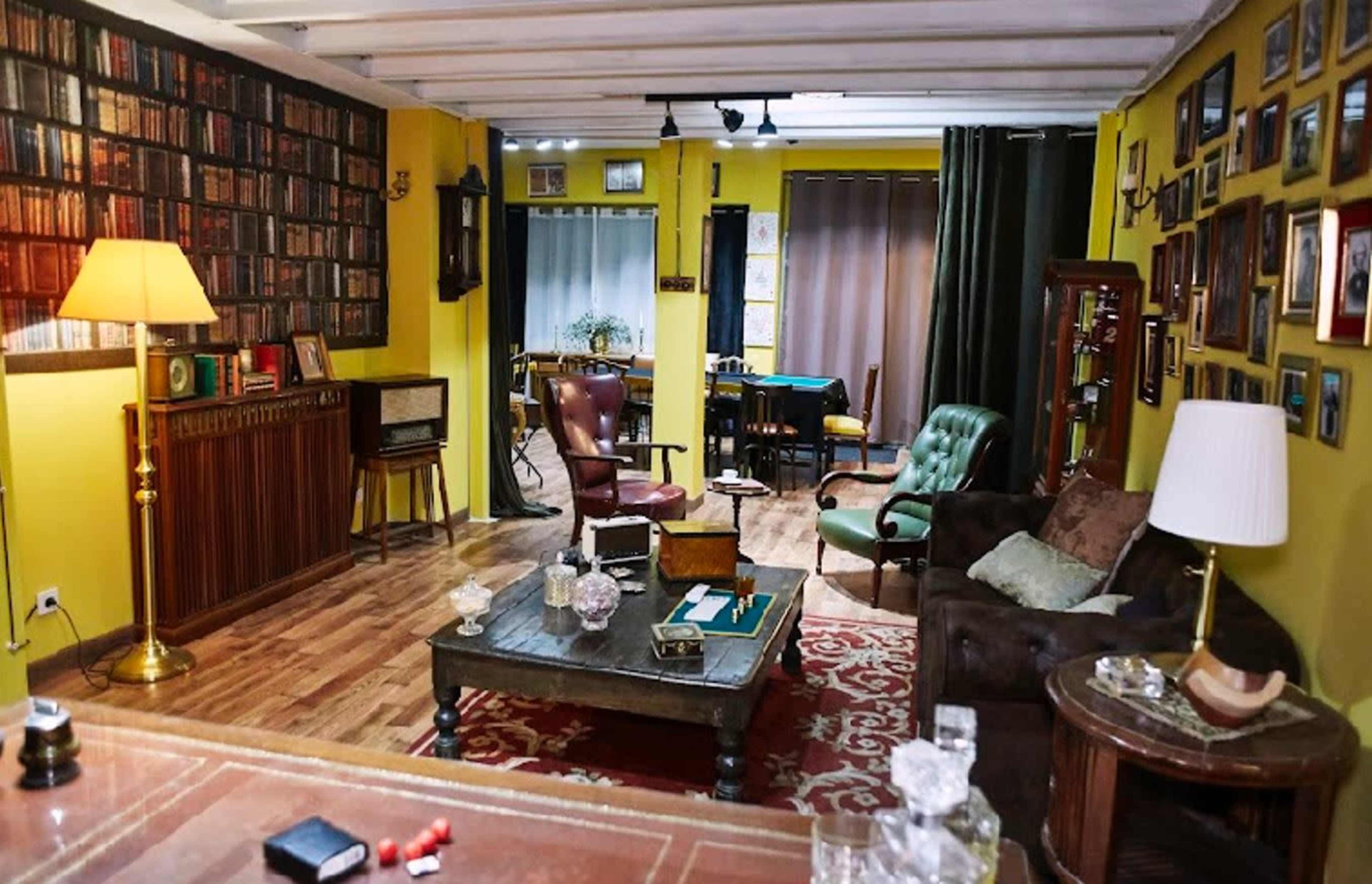 The room features a cozy living space with wooden flooring, a mix of vintage furniture, framed photographs on the walls, and a warm lamp illuminating the area.