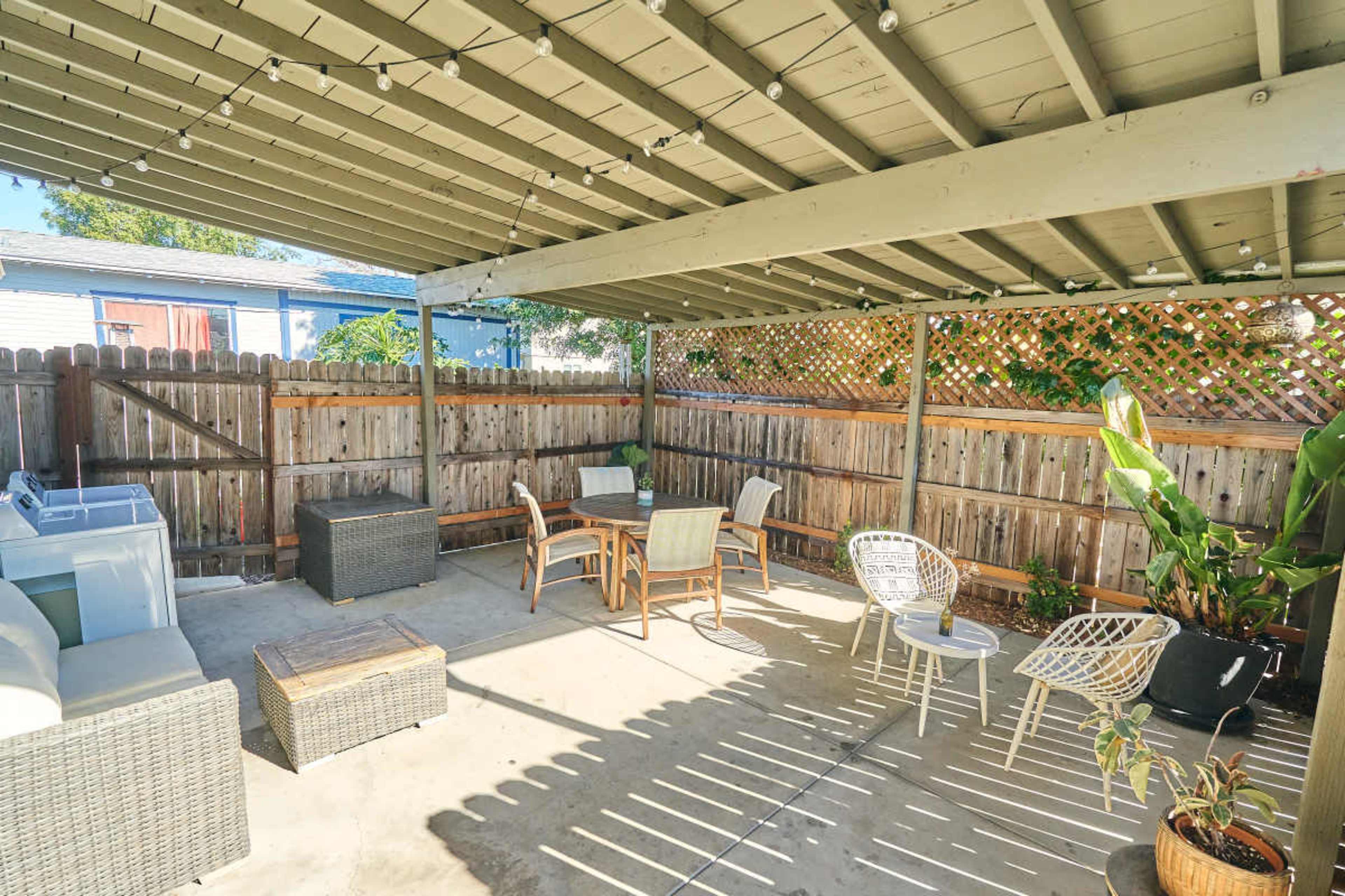 The image shows a covered outdoor patio area with several seating options, a small table, and a laundry area against a wooden fence.