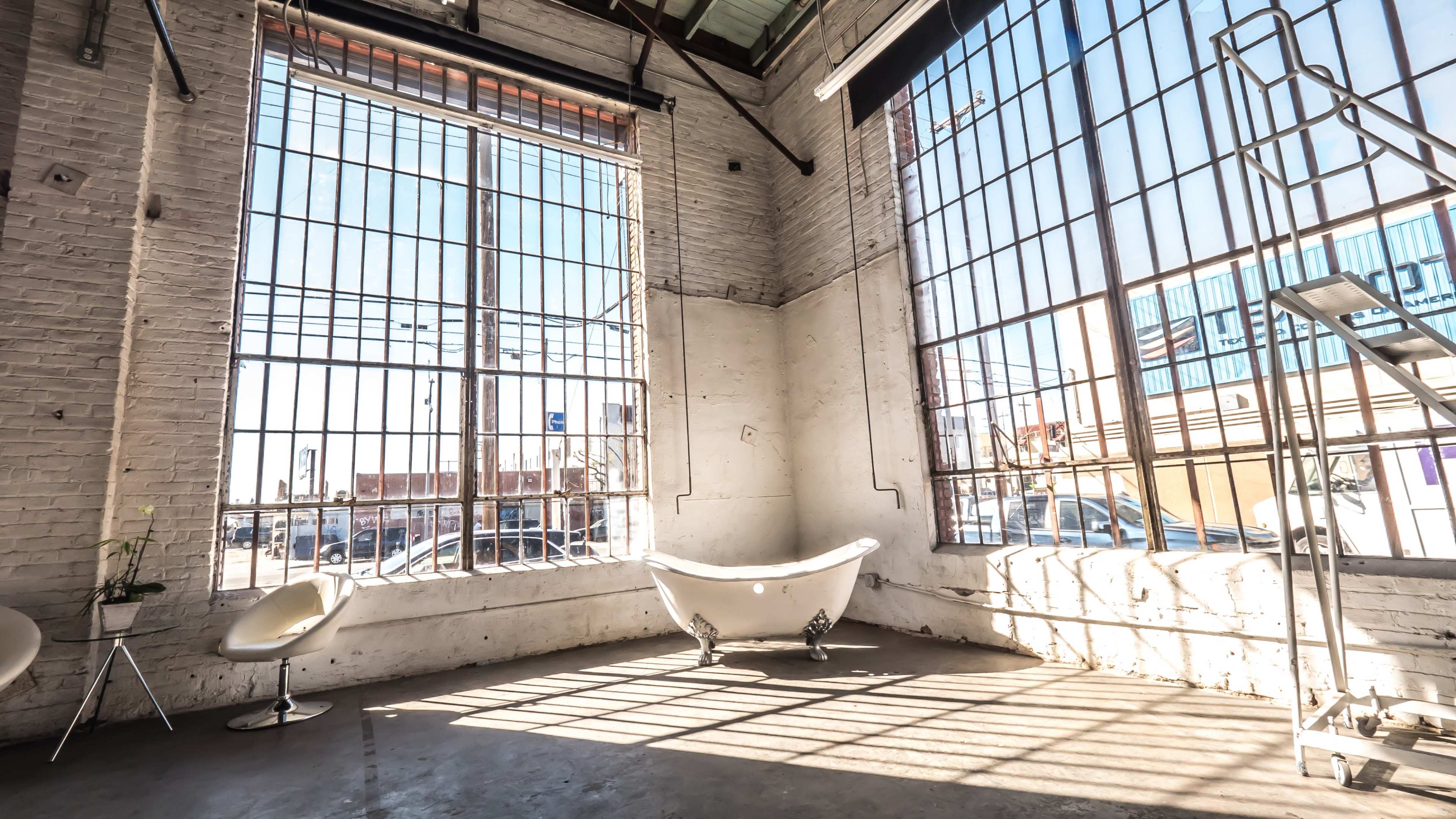 The image shows a spacious, bright room with large windows, featuring a freestanding bathtub and two modern chairs.