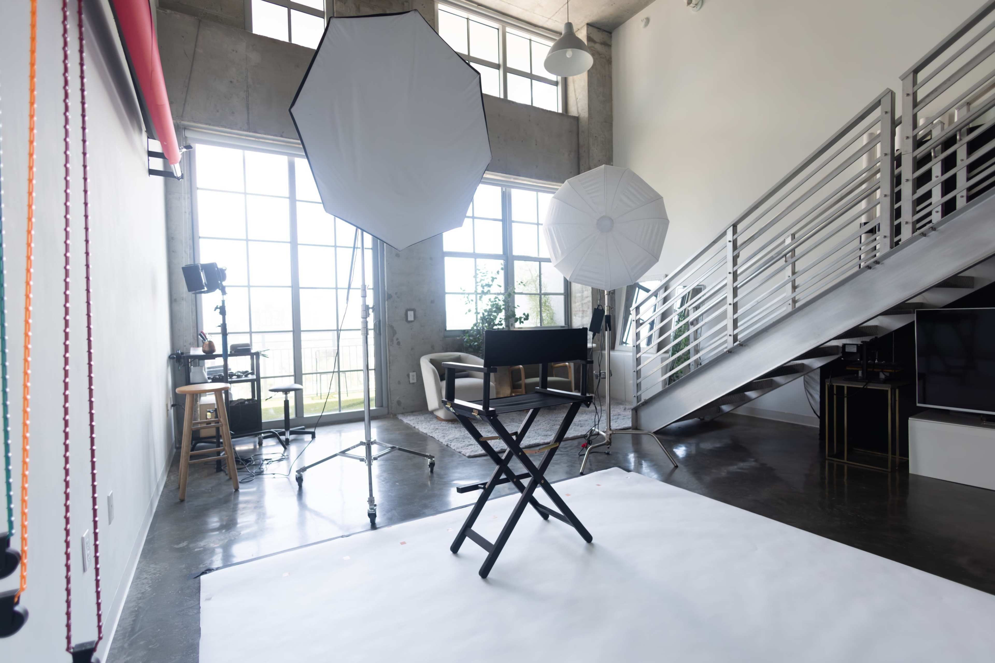 The image shows a spacious photography studio featuring large windows, two light umbrellas, a black director's chair on a white backdrop, and a staircase leading to an upper level.