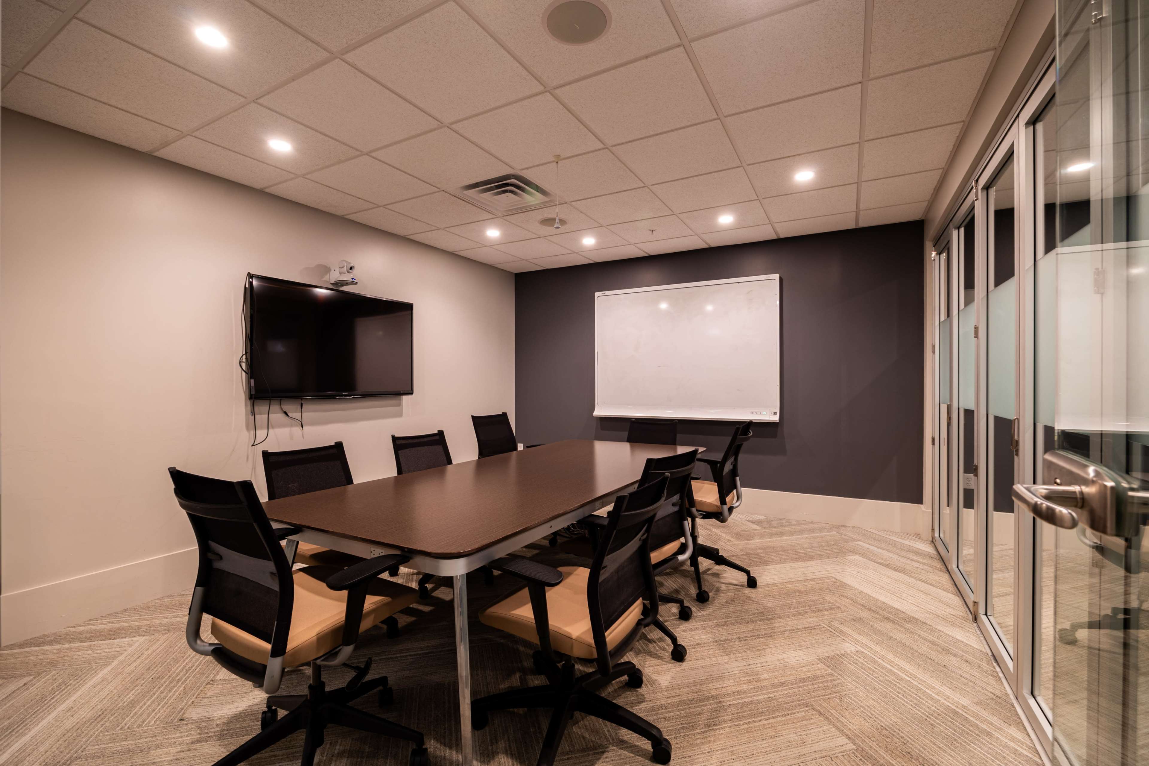 A conference room features a long table surrounded by chairs, a wall-mounted television, and a whiteboard.