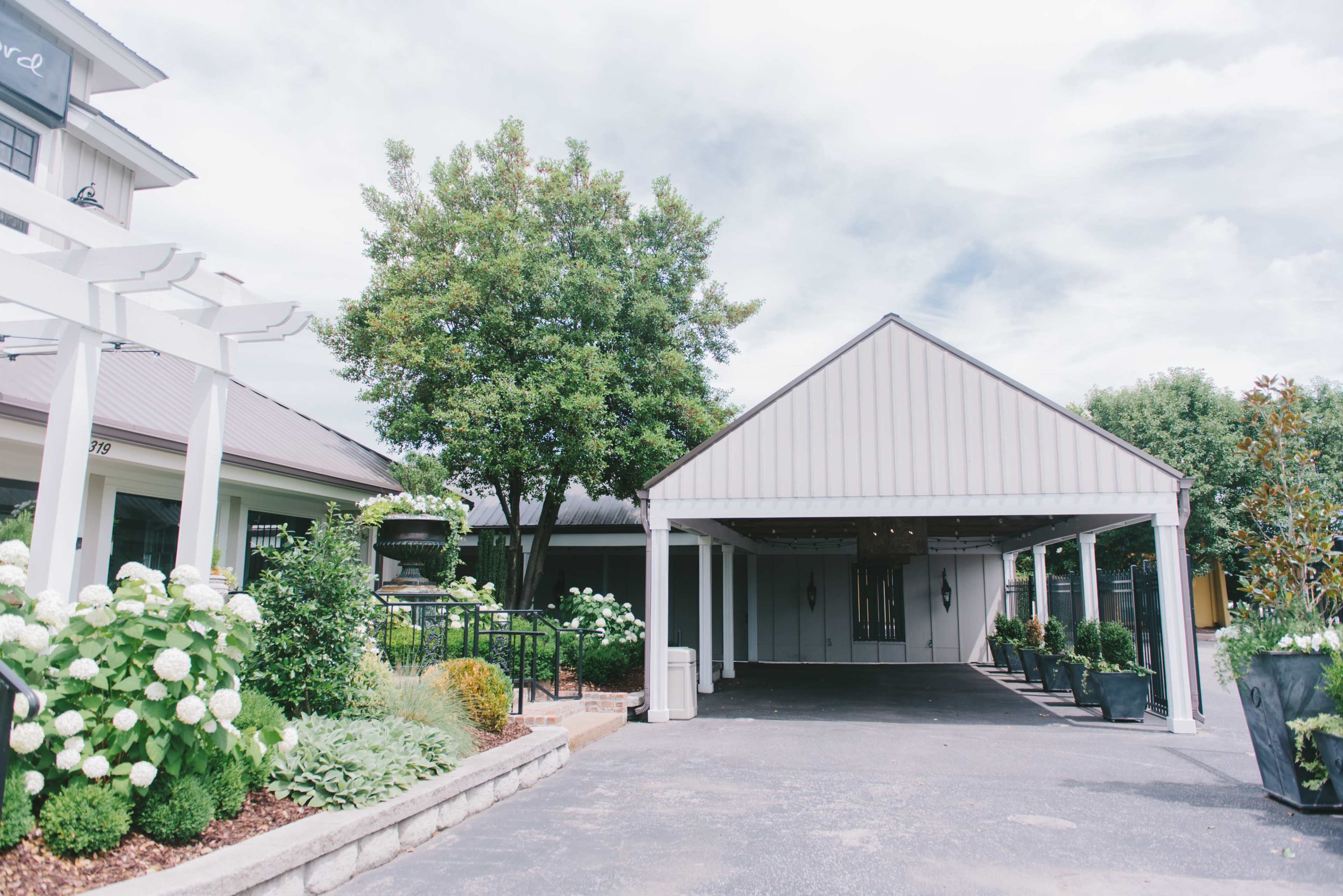 A driveway leading to a modern building with a covered entrance, surrounded by landscaped greenery and flower beds.