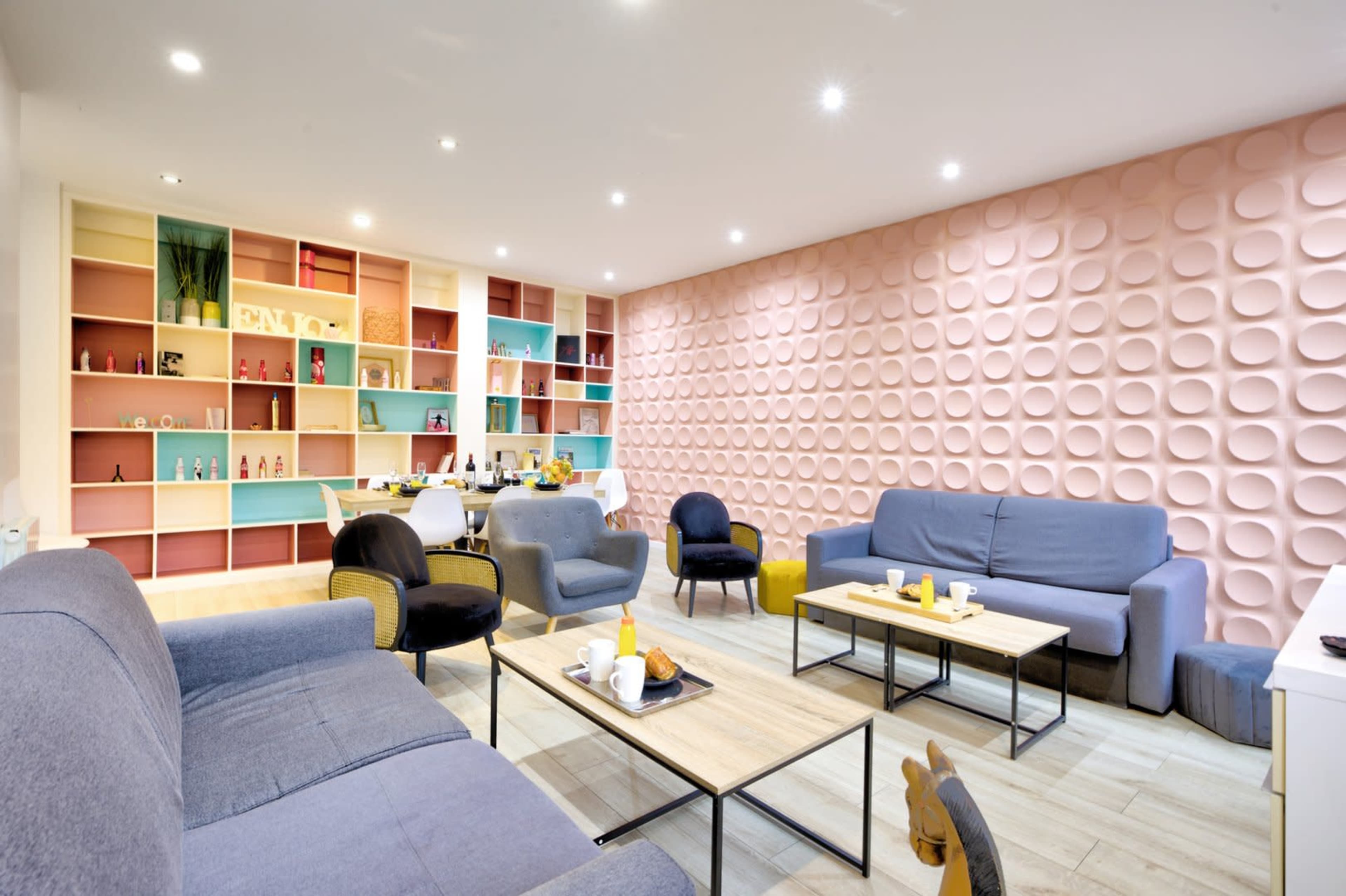 A modern lounge area features blue and black sofas, wooden tables, and a colorful shelving unit against a textured pink wall.