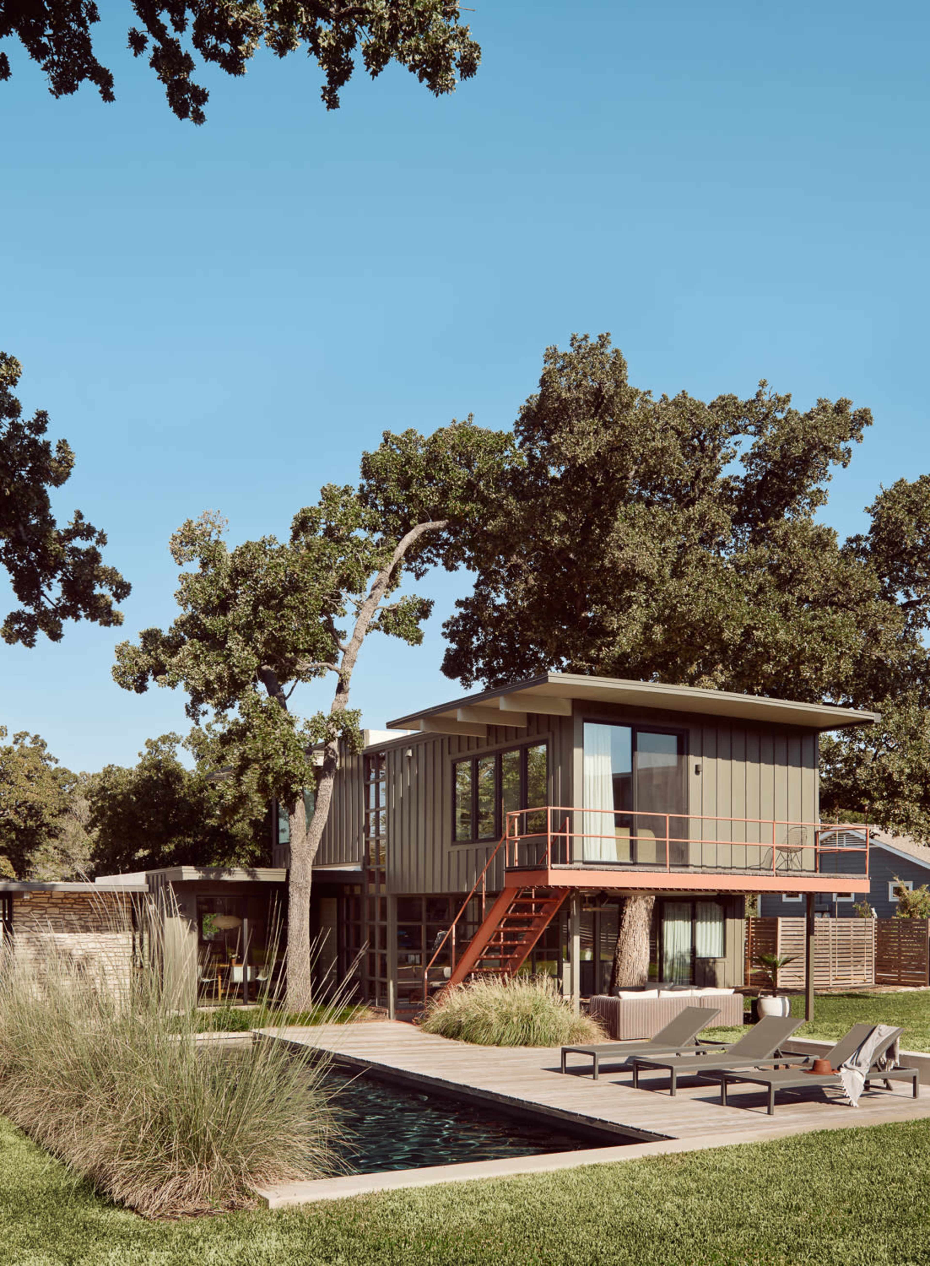 A modern two-story house is surrounded by greenery, featuring large windows, a red staircase, and a swimming pool in the foreground.