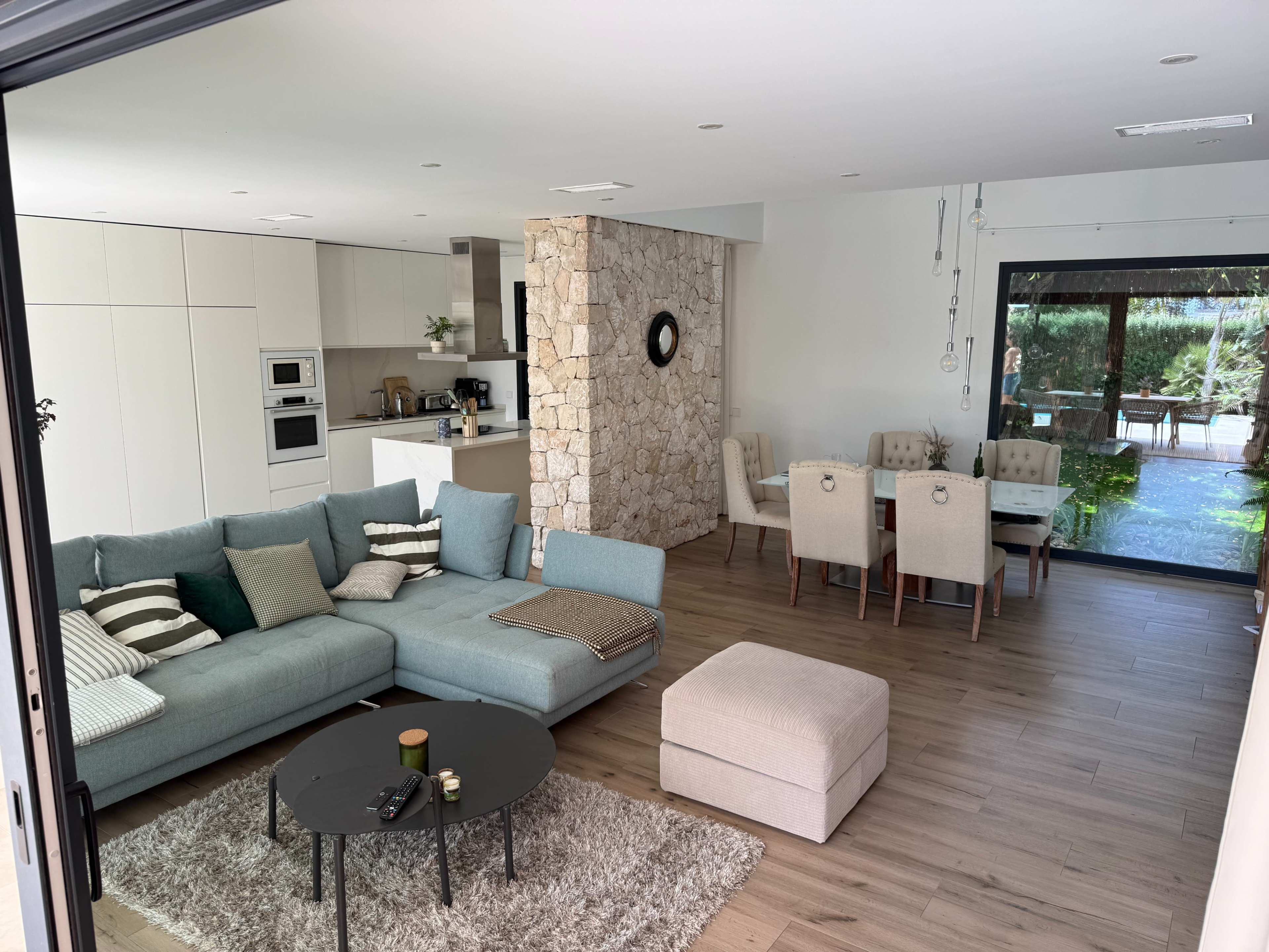 The image shows a modern living room with a large sectional sofa, a coffee table, and a dining area featuring a stone wall and large windows.