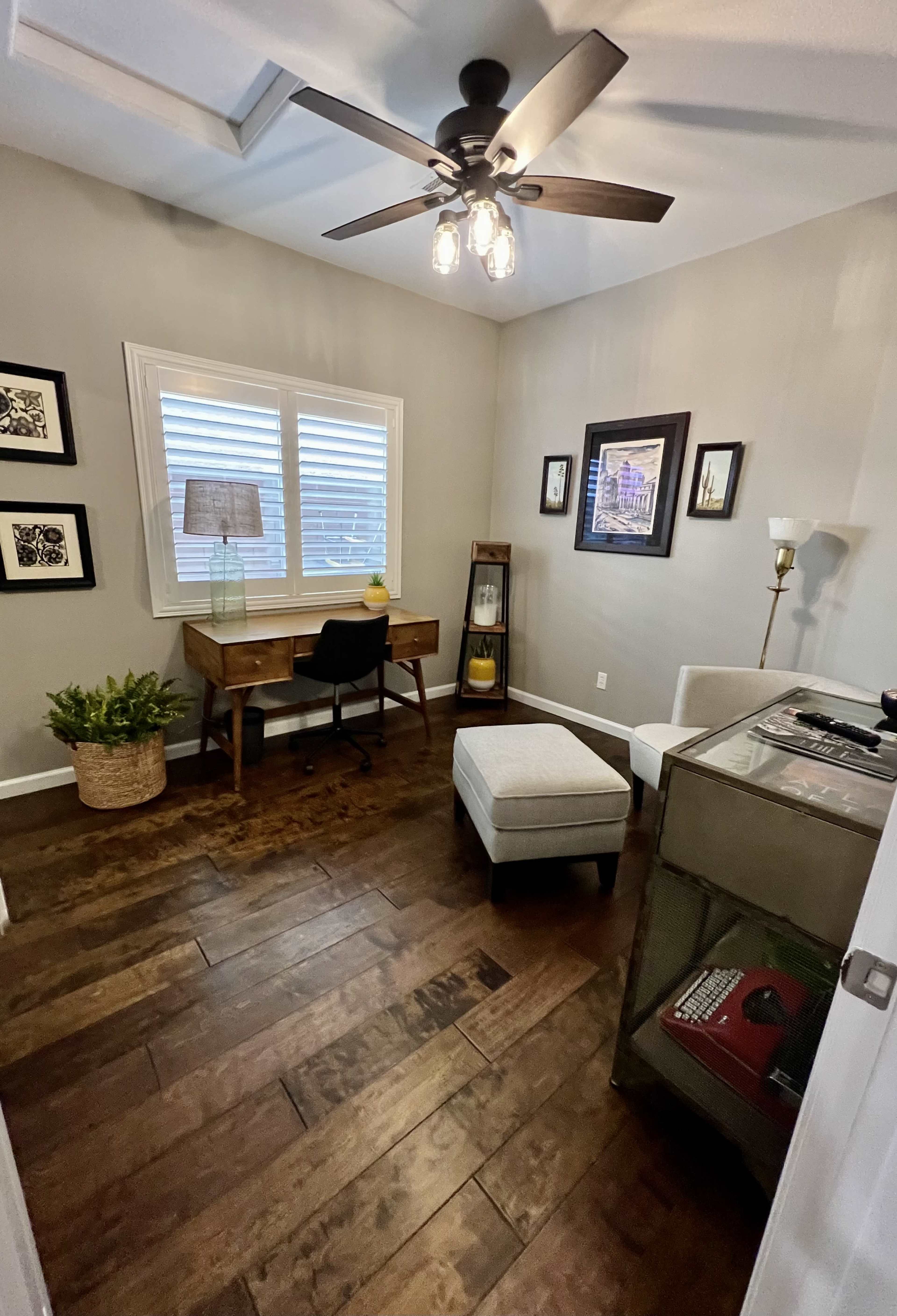 The image shows a small, well-lit home office featuring a wooden desk, a chair, a lamp, a potted plant, and a display cabinet against a wall.