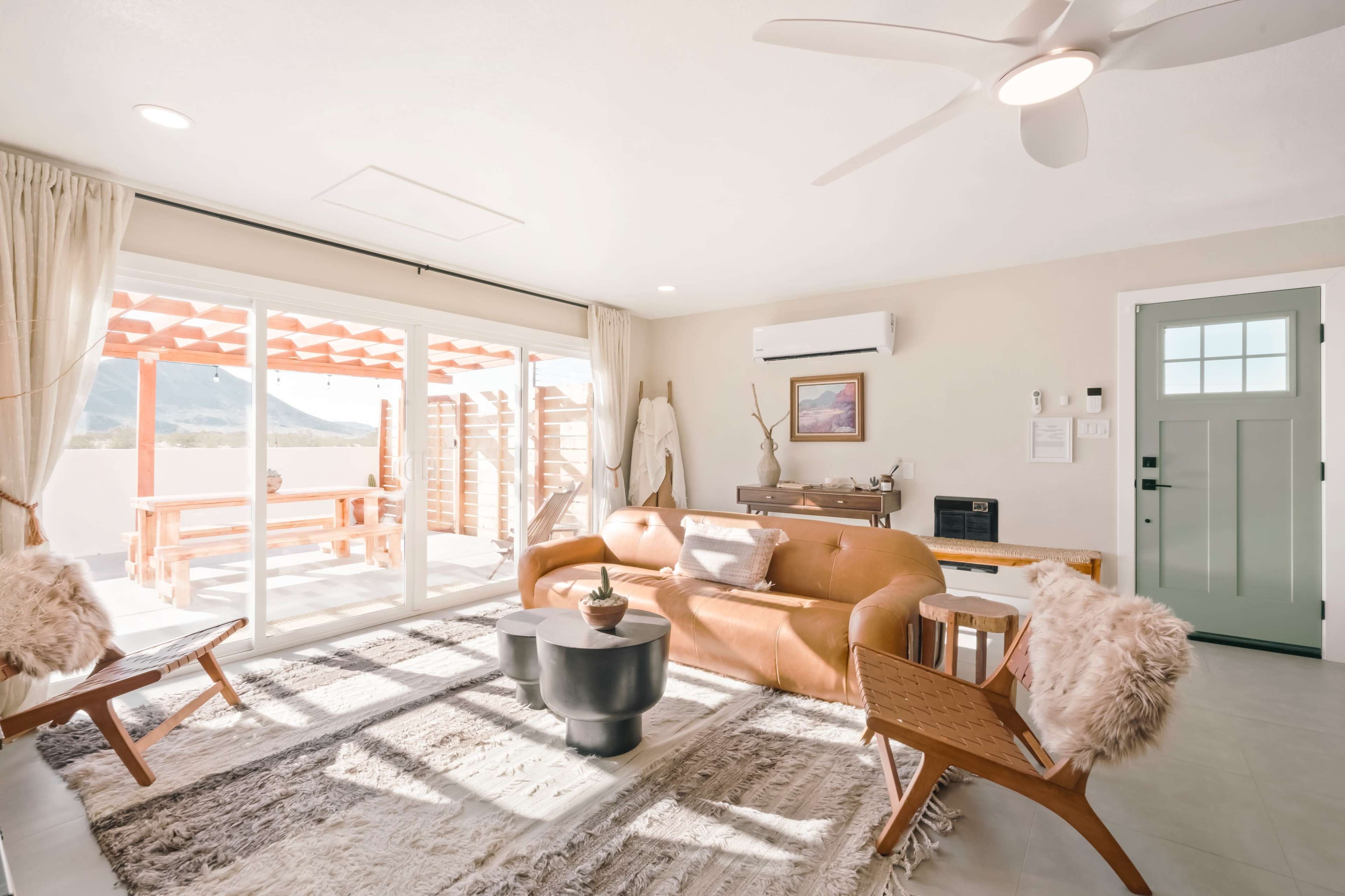 A bright, modern living room with a brown leather sofa, wooden chairs, and large sliding glass doors leading to an outdoor area.