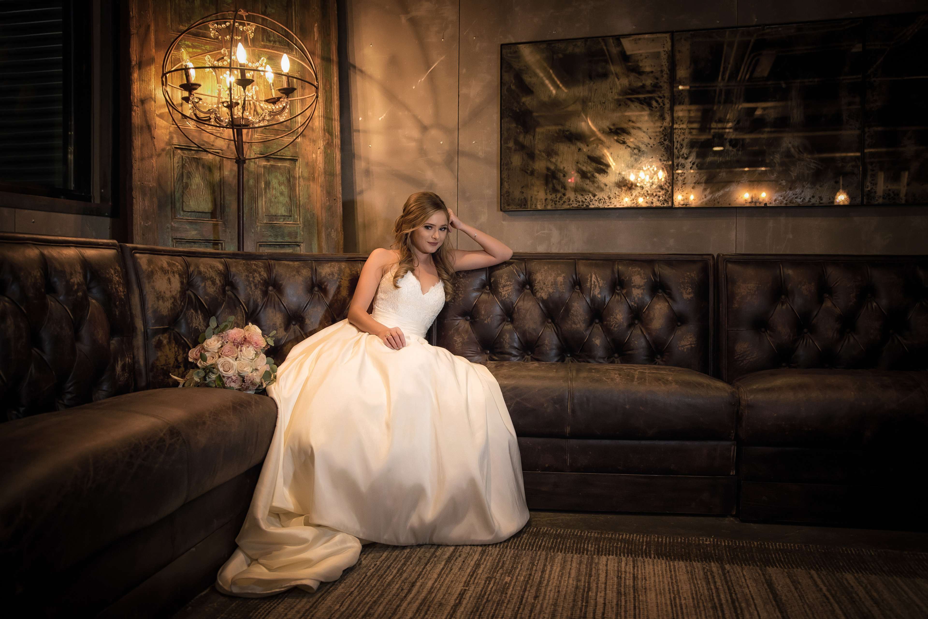 A bride in a white gown sits elegantly on a brown leather couch in a dimly lit room, holding a bouquet of flowers.