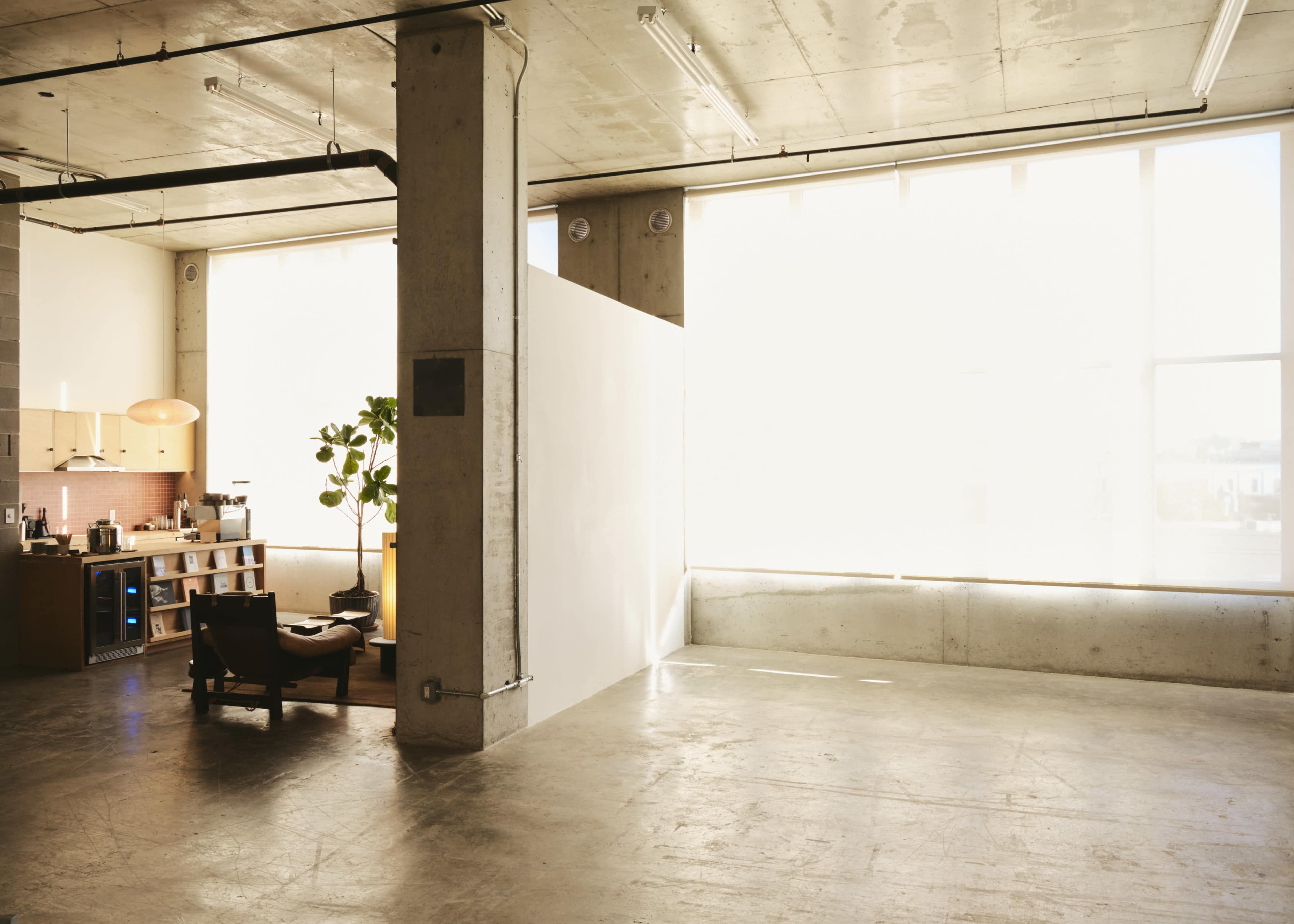 A spacious and minimally furnished industrial-style loft features large windows, concrete walls, and a small kitchen area with a dining space.