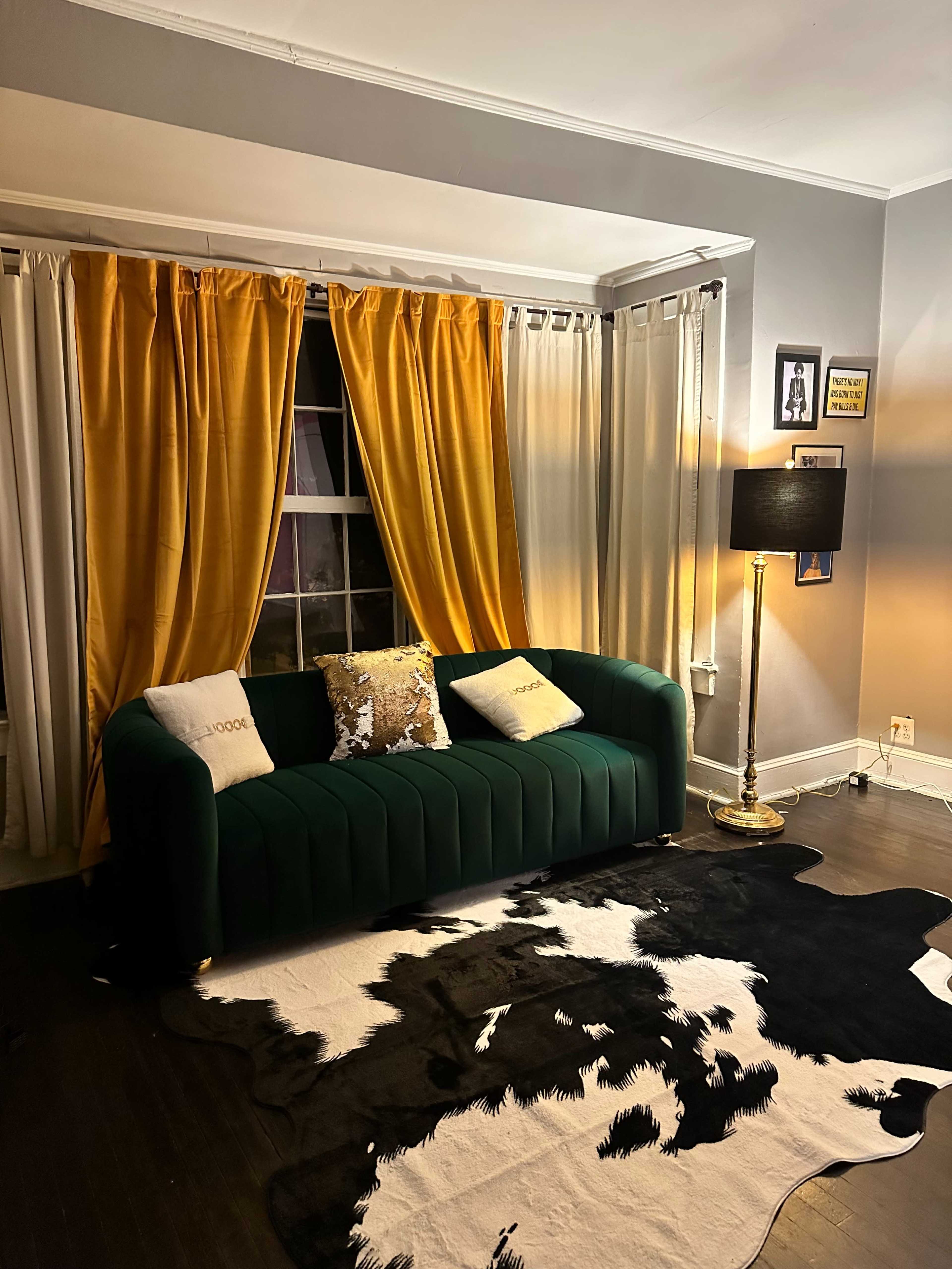 A green sofa is positioned in a living room with yellow and white curtains, a gold floor lamp, and a cowhide rug on a dark wood floor.