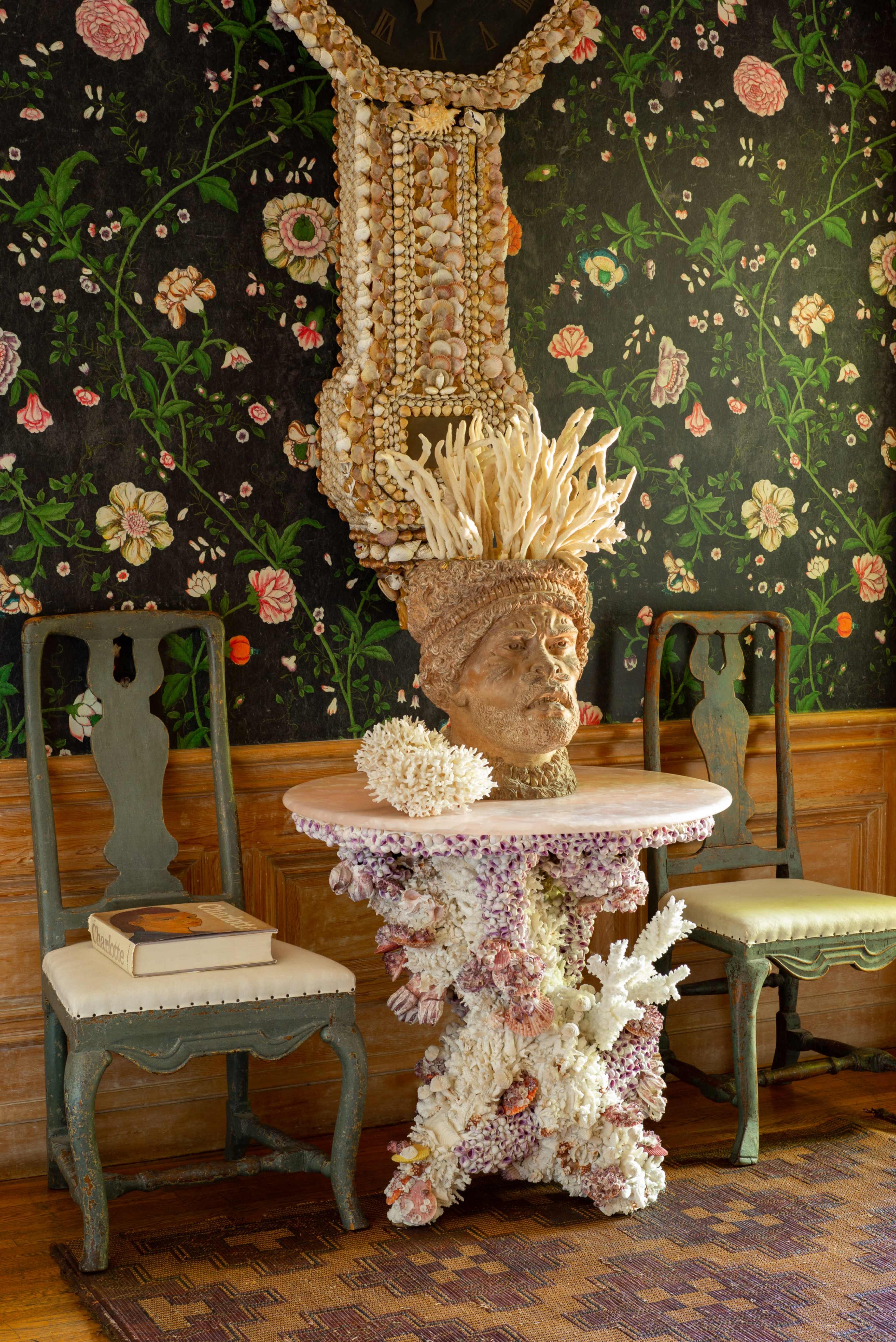 The image shows a decorative corner of a room featuring a coral-inspired table with a bust on top, two vintage chairs, and a floral-patterned wallpaper.