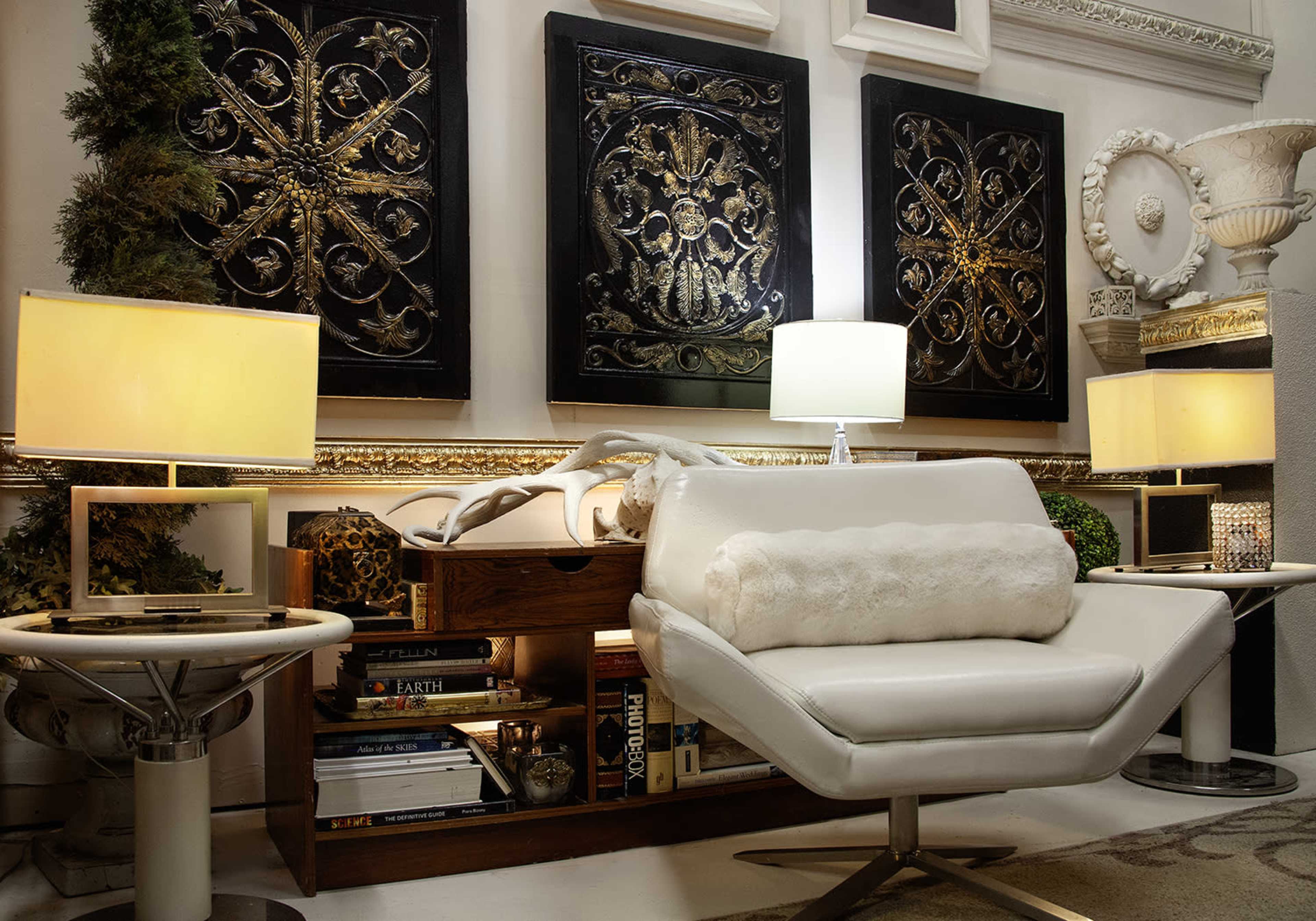 The image shows a modern living space featuring a white chair with a fur accent, surrounded by decorative wall art, lamps, and books neatly arranged on a shelf.