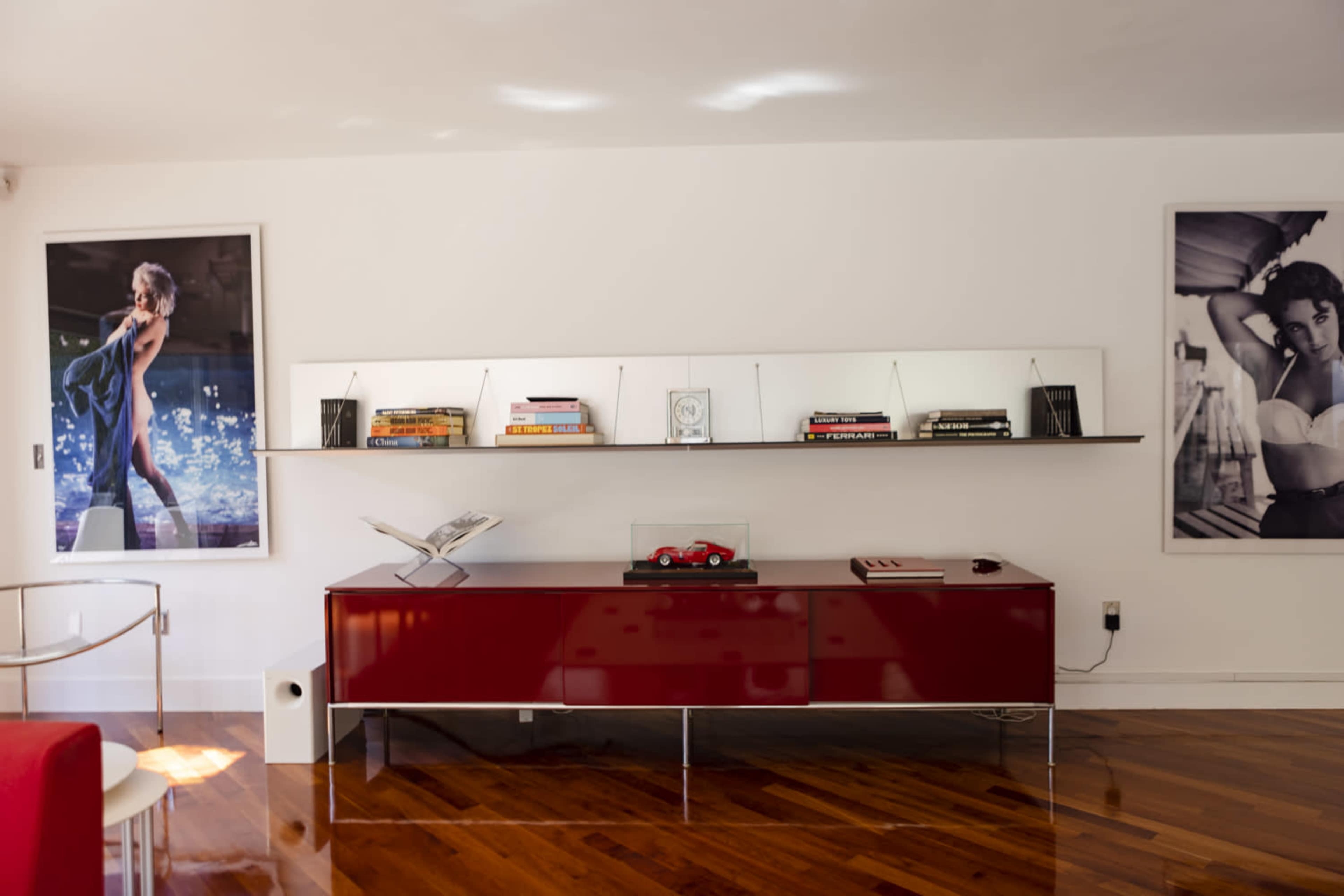 The image shows a modern living room with a red low cabinet beneath two large framed photographs and a shelf displaying books and decor.