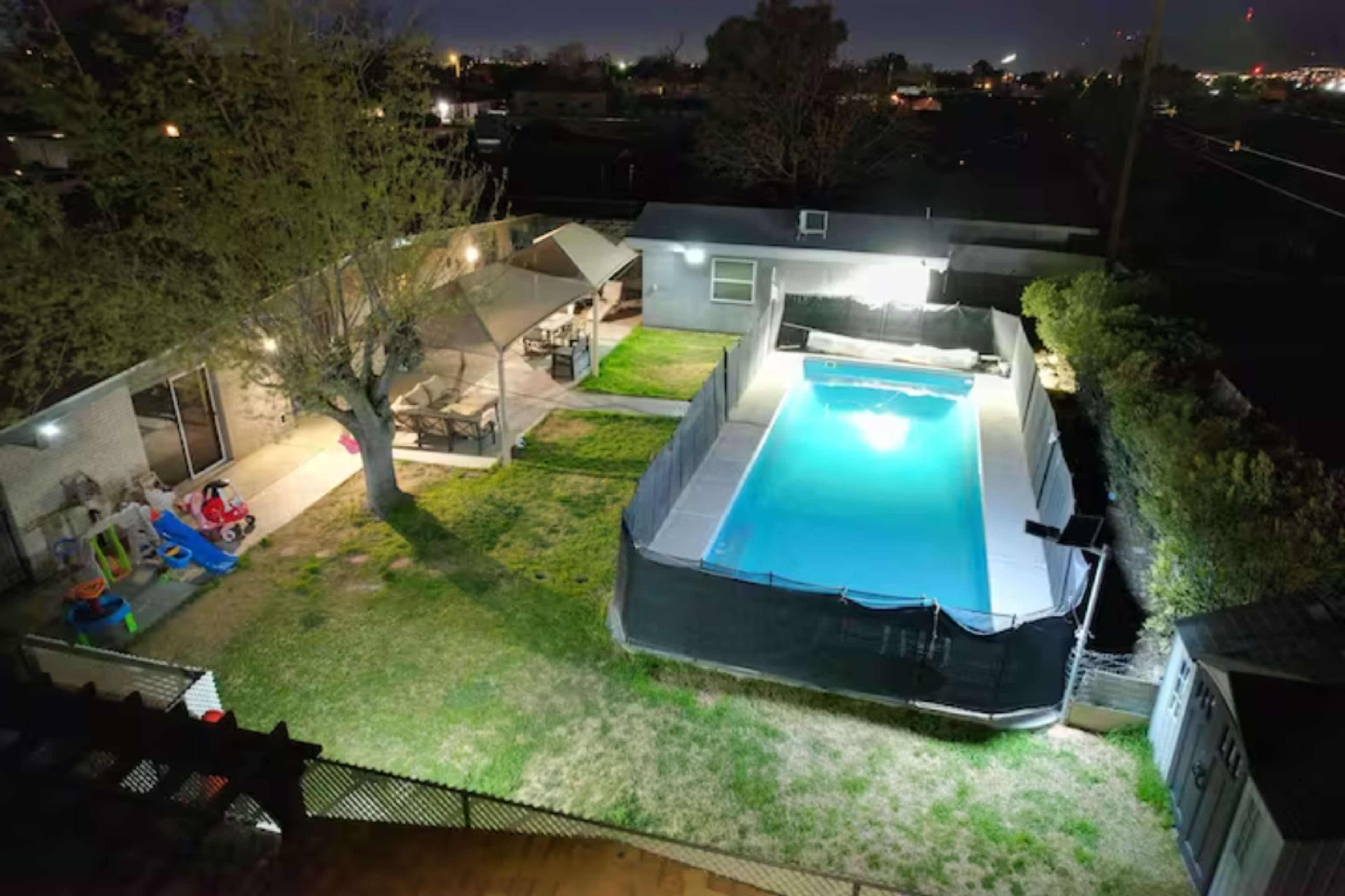 The image shows a backyard scene at night featuring a swimming pool surrounded by grass, with a seating area and play equipment nearby.
