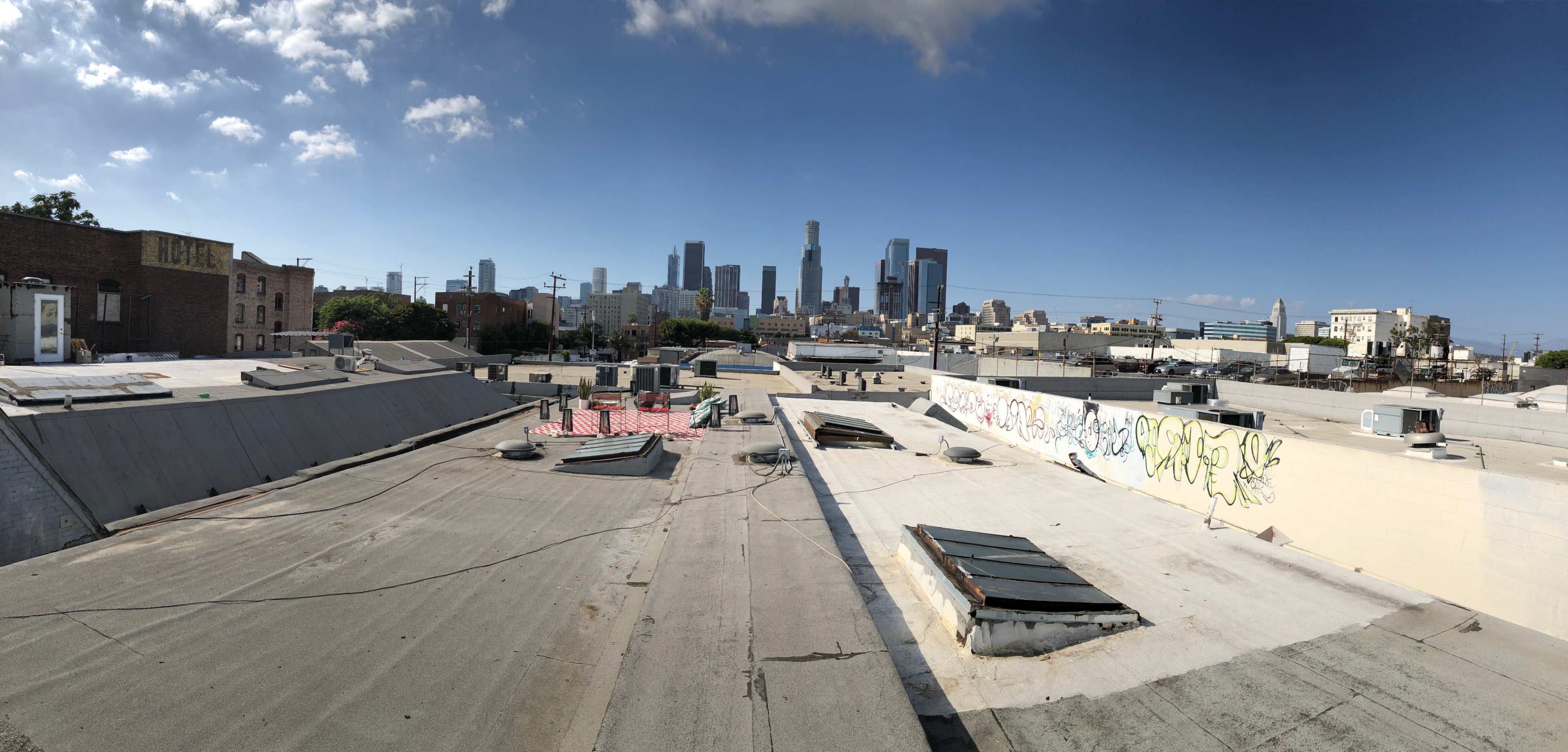A panoramic view of rooftops in the foreground with the skyline of downtown Los Angeles in the background under a partly cloudy sky.