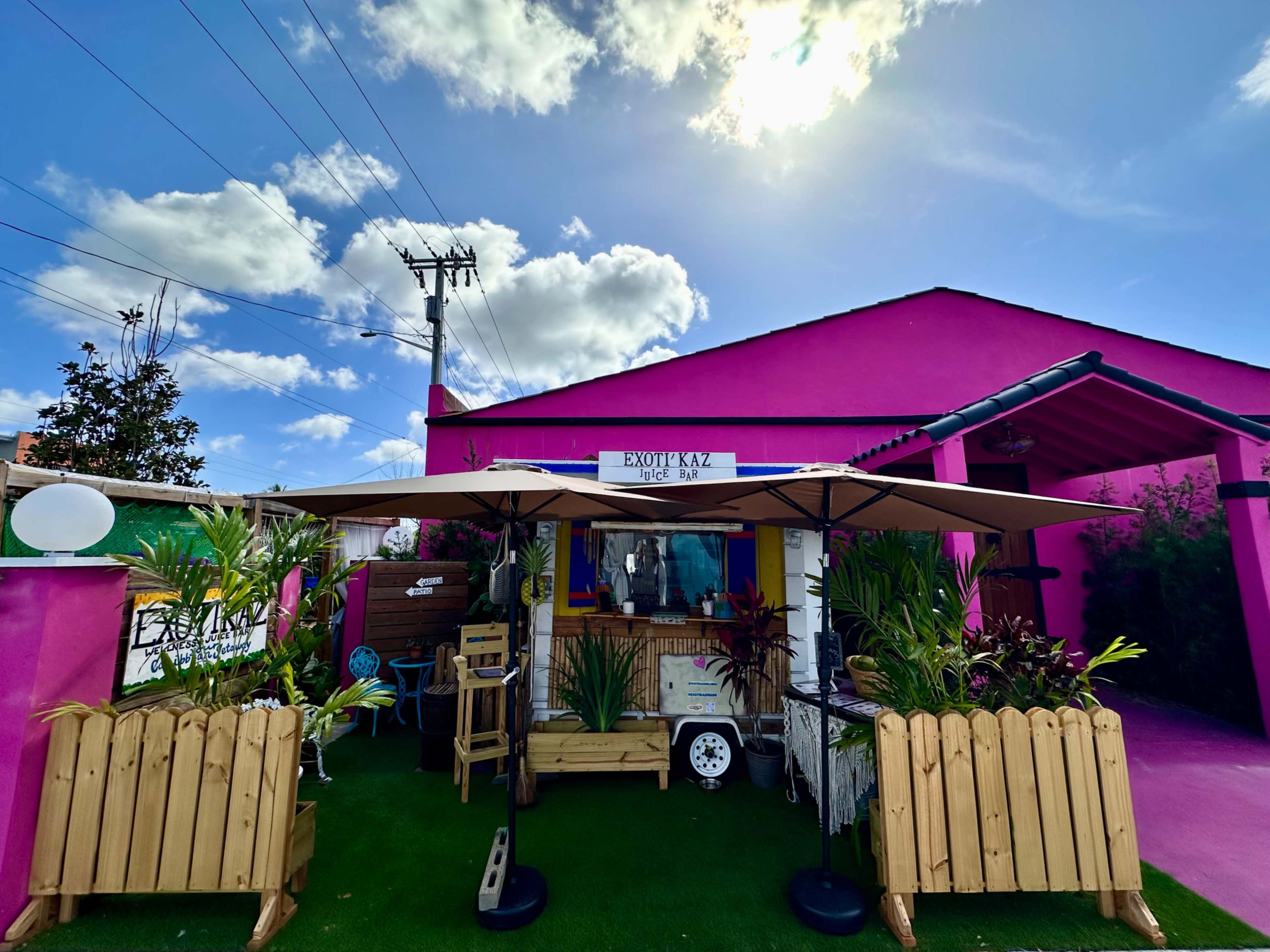The image shows a vibrant pink building with outdoor seating areas and umbrellas surrounded by greenery and decorative elements.