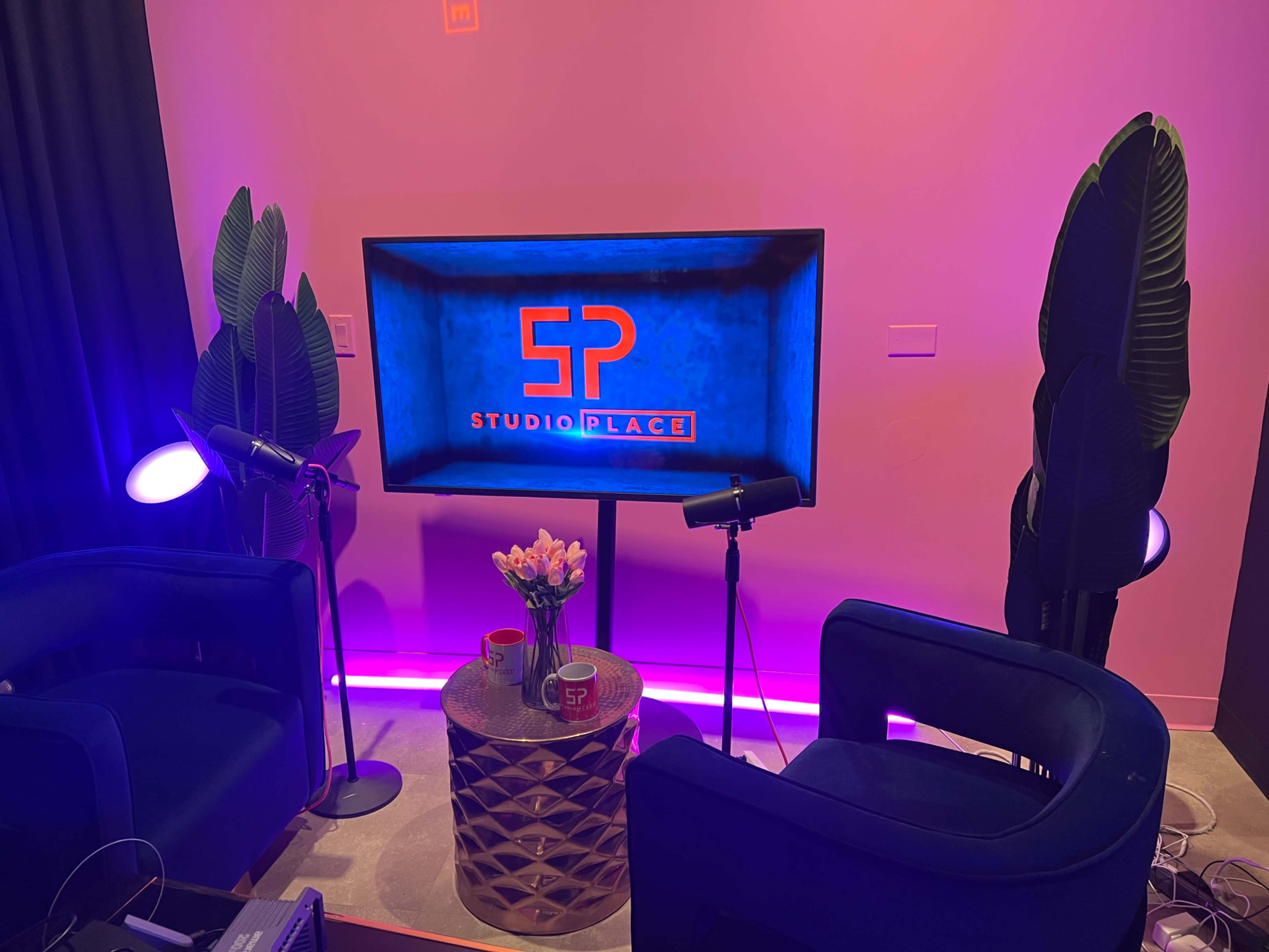 A modern studio setup with two blue chairs, a round table with flowers, and a television displaying the logo "5P STUDIO PLACE" against a pink wall.