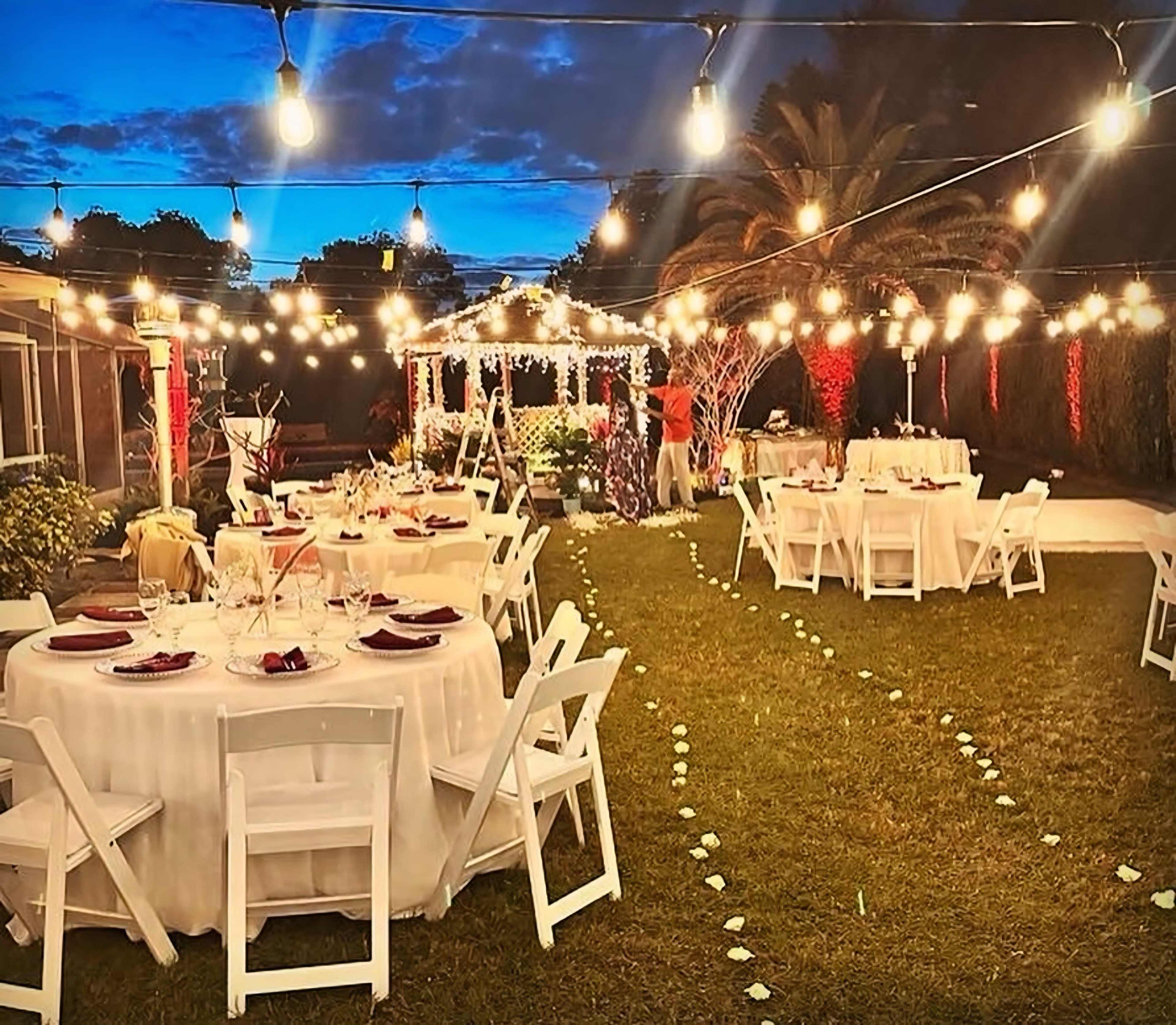 The scene depicts an outdoor garden setting for a festive event, with elegantly arranged tables and string lights illuminating the area.