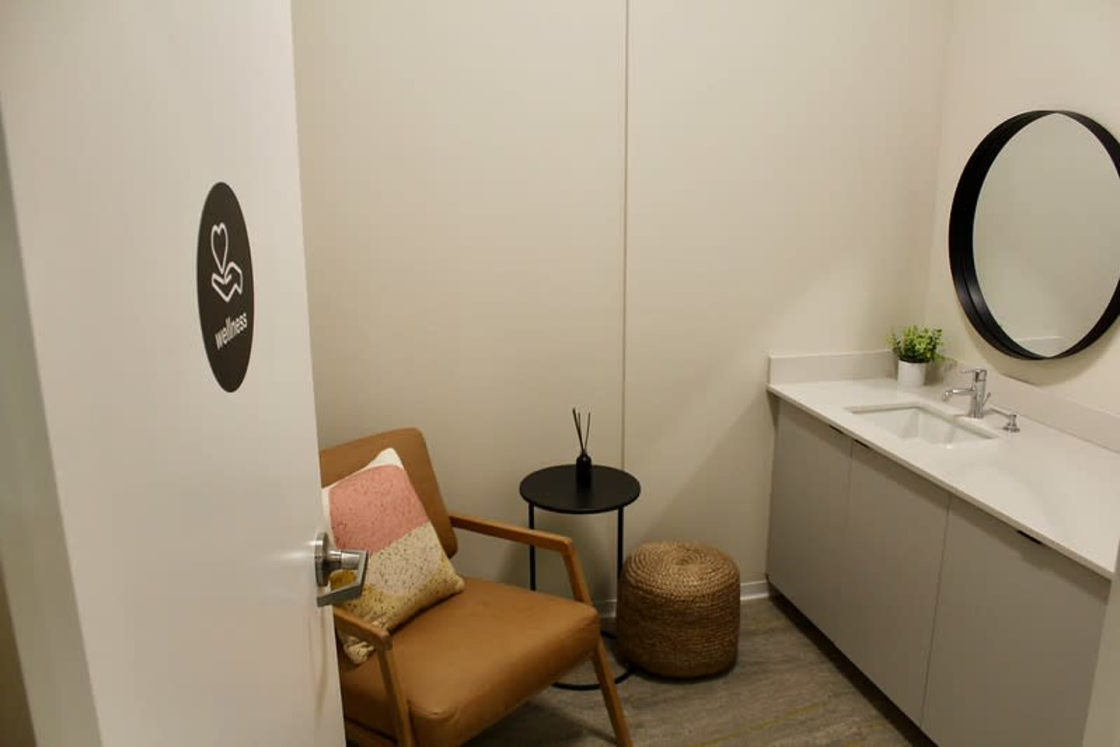 The image shows a small wellness room with a chair, a round mirror, a sink, and a small table, featuring neutral-colored walls and flooring.