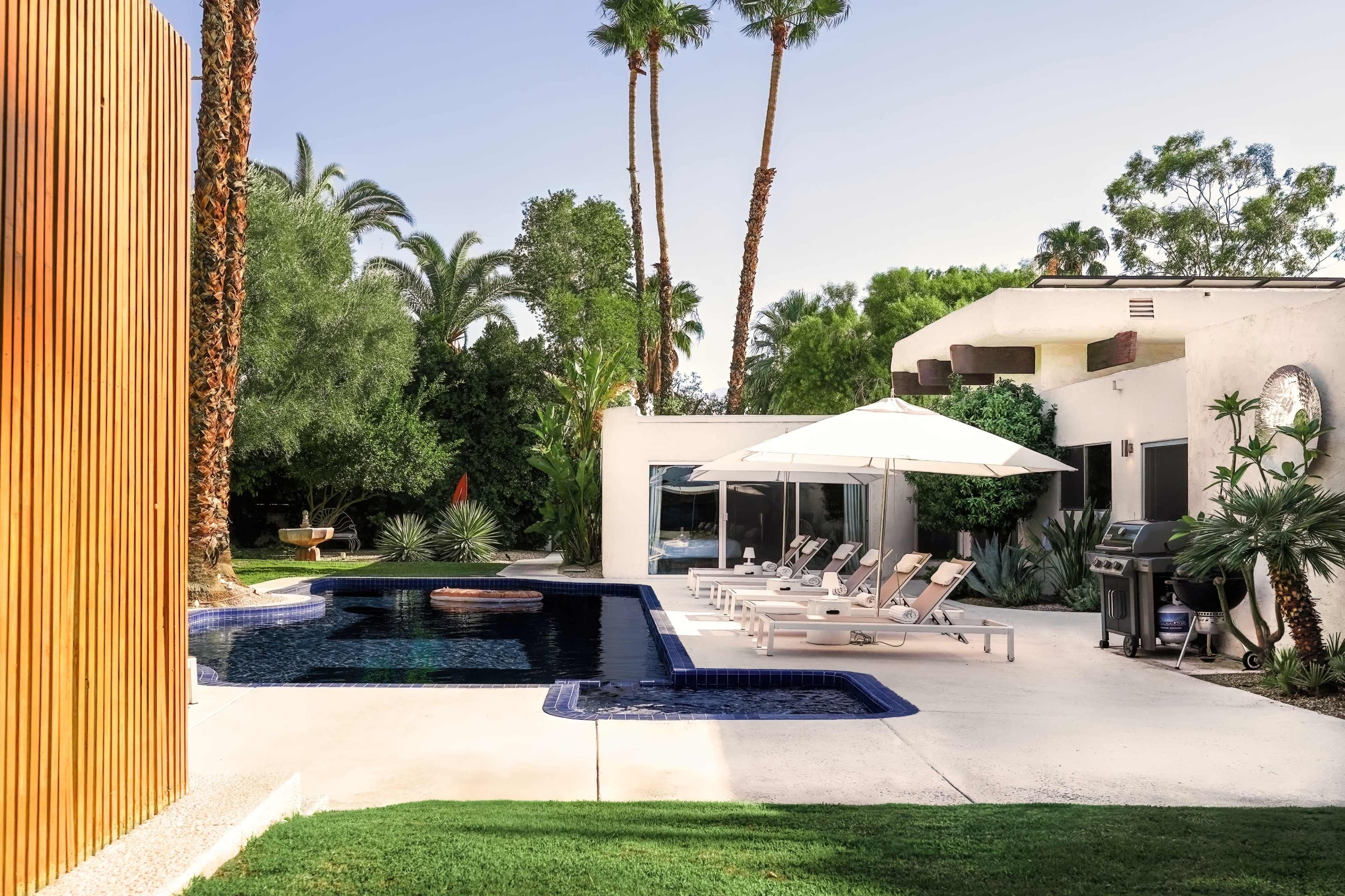 A modern backyard with a swimming pool, lounge chairs under an umbrella, and a barbecue area, surrounded by tropical landscaping and palm trees.