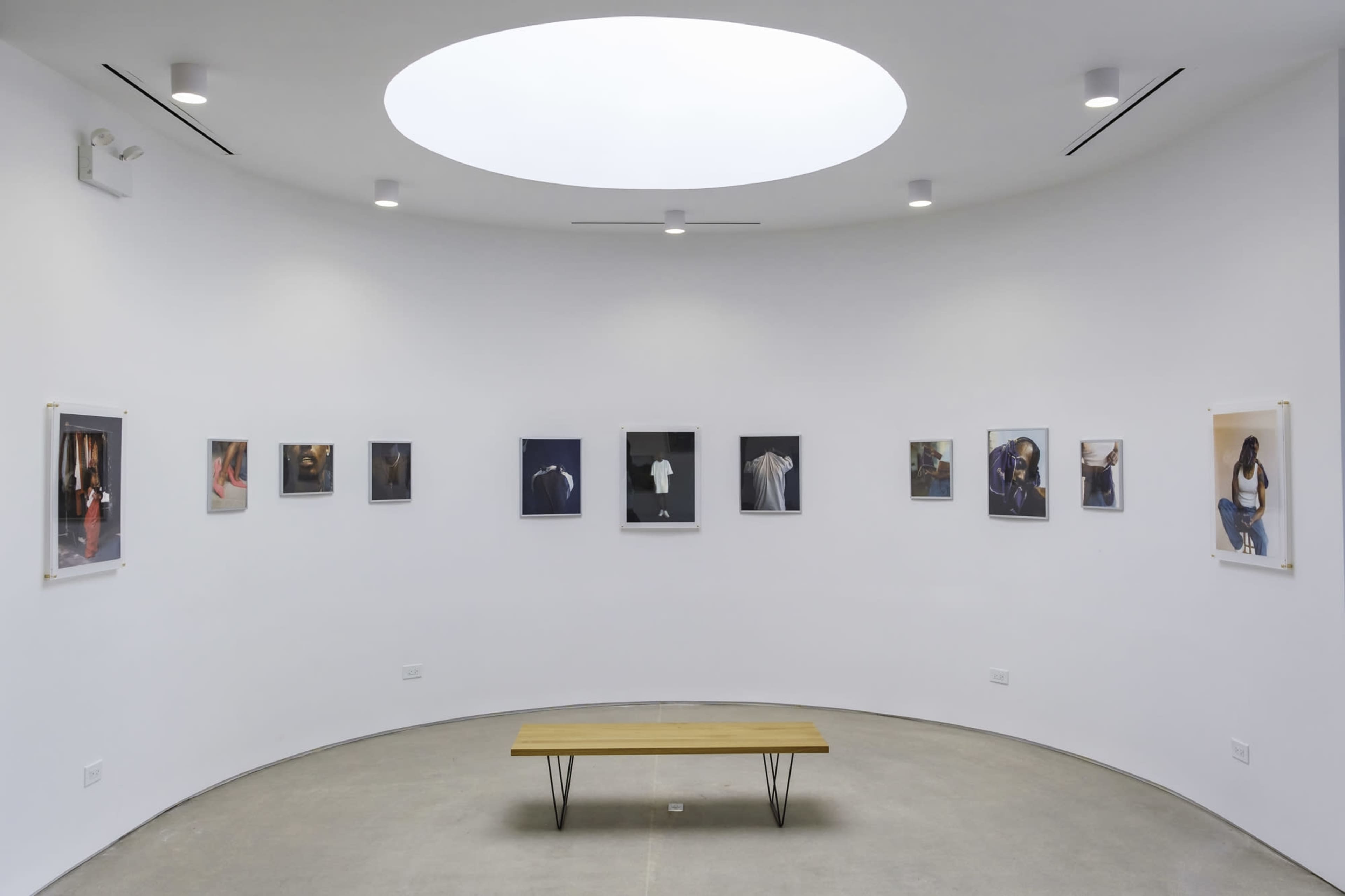 The image shows a circular gallery space with a skylight, featuring several framed photographs hung on the walls and a wooden bench in the center.