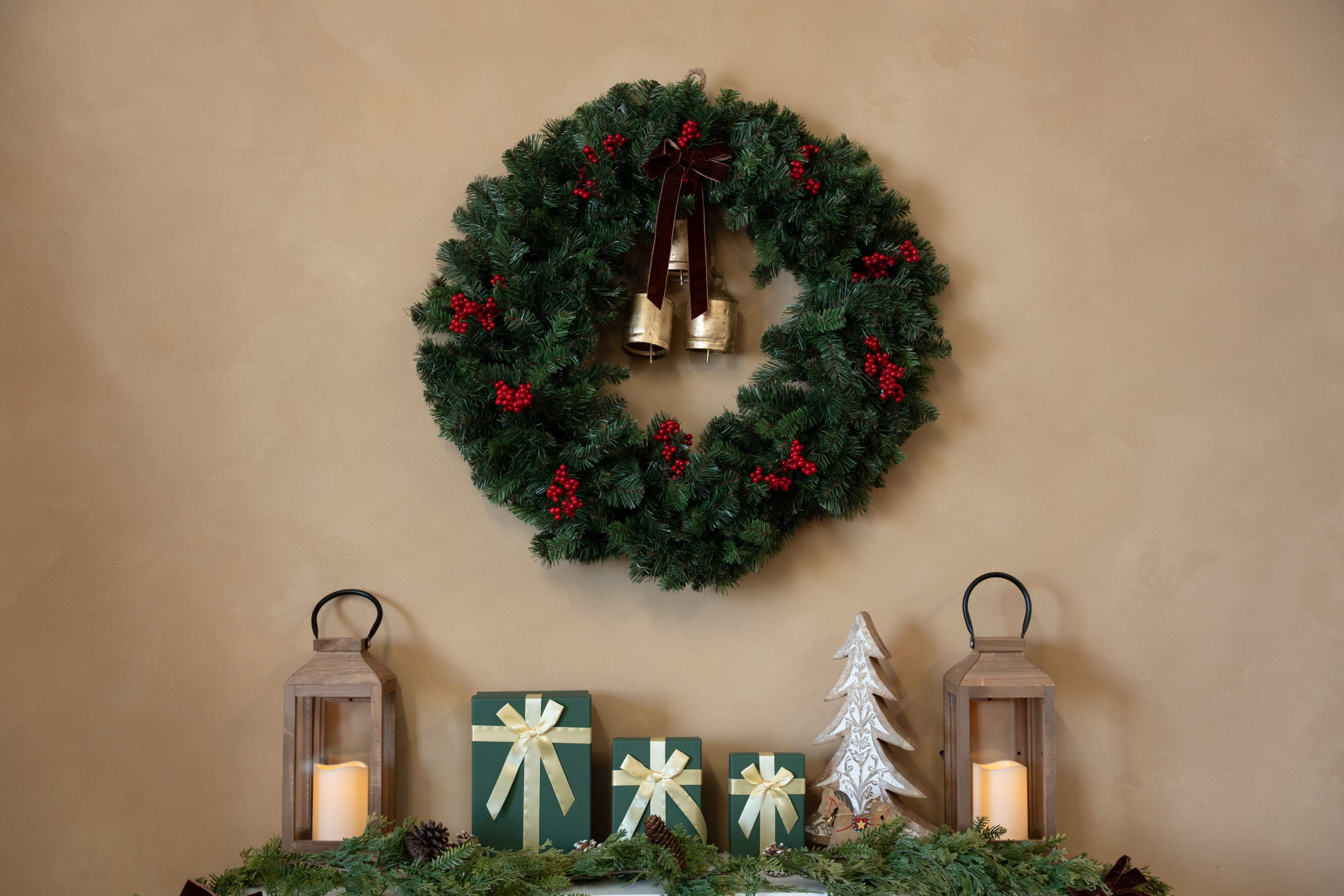 A festive wreath decorated with holly and bells hangs on a wall above a table adorned with wrapped gifts and lanterns.