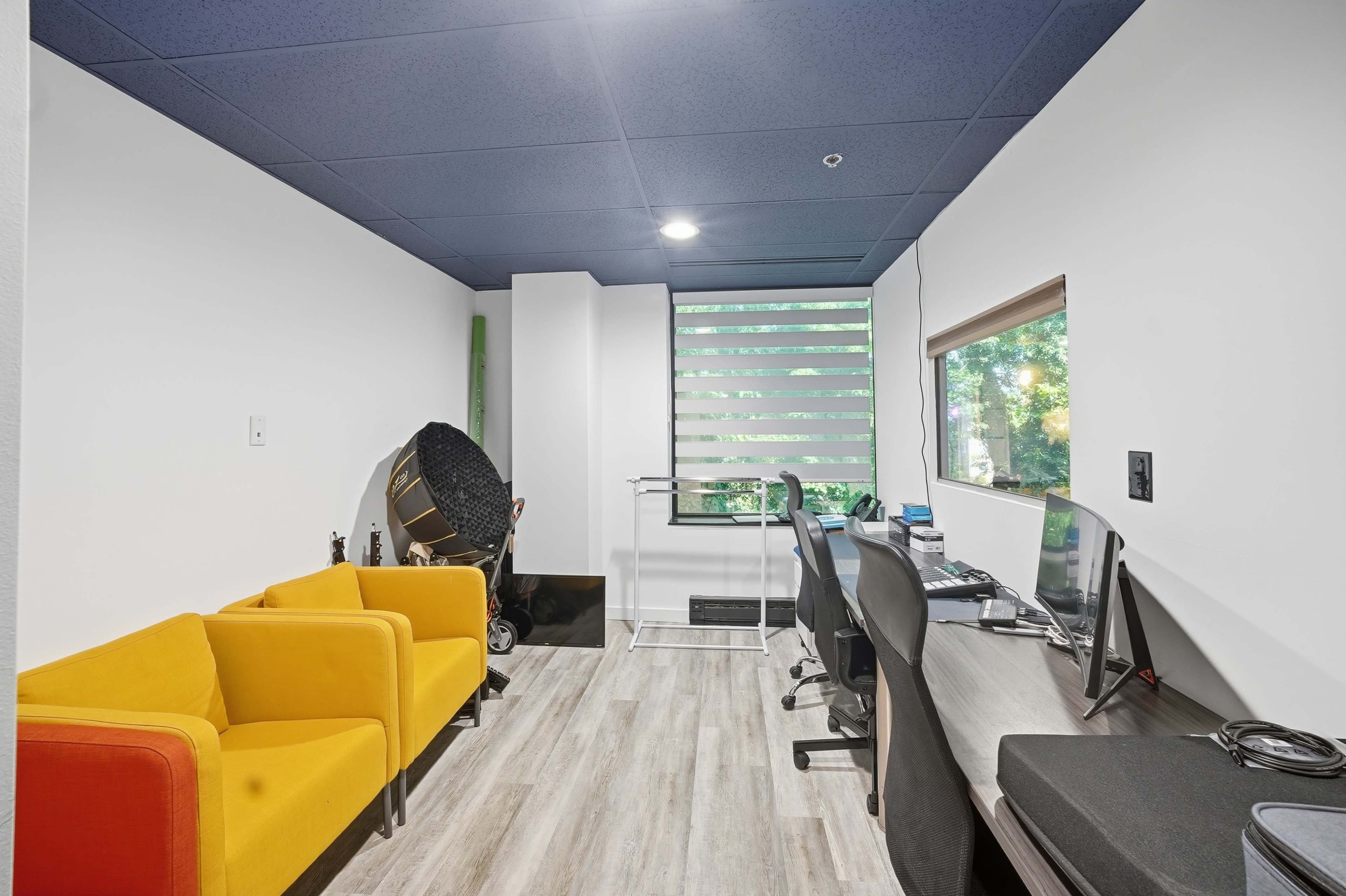 The image shows a small office space with two desks, a yellow couch, and large windows with blinds, allowing natural light to enter.