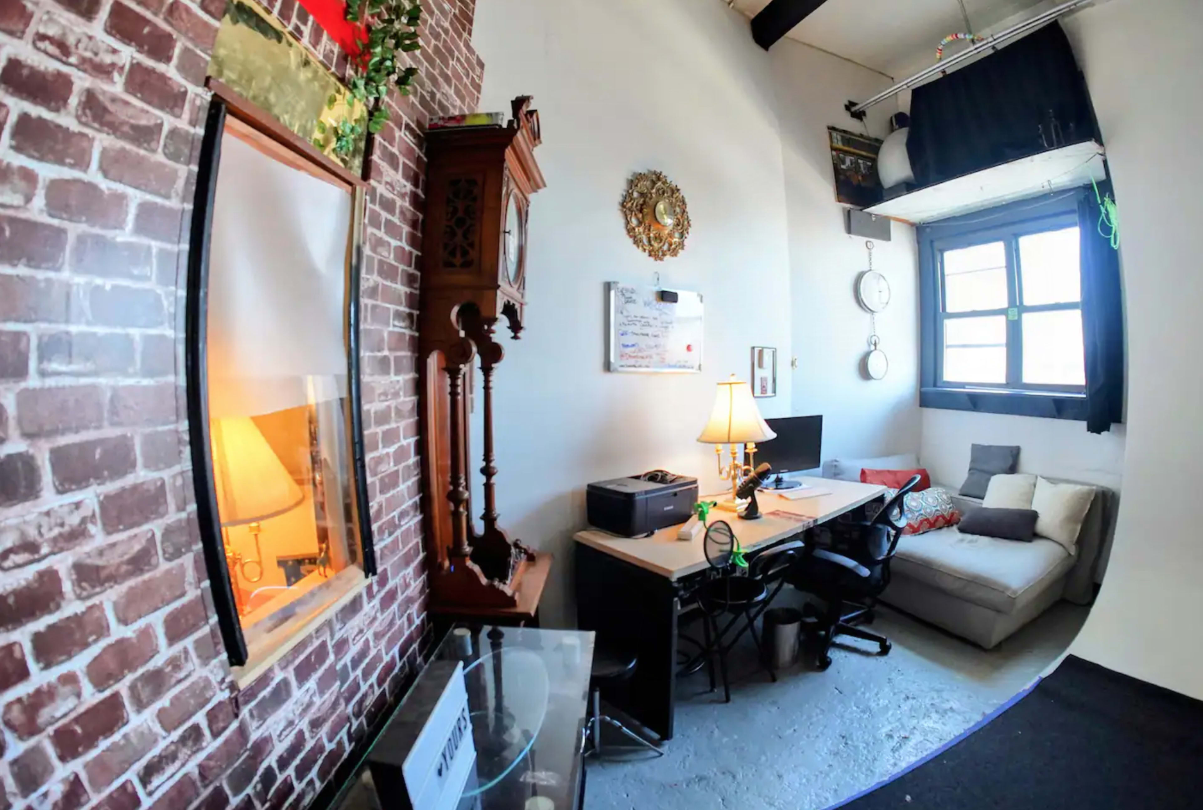 A cozy home office with a brick wall, a desk with a computer and printer, a large clock, and a sofa in a well-lit corner.
