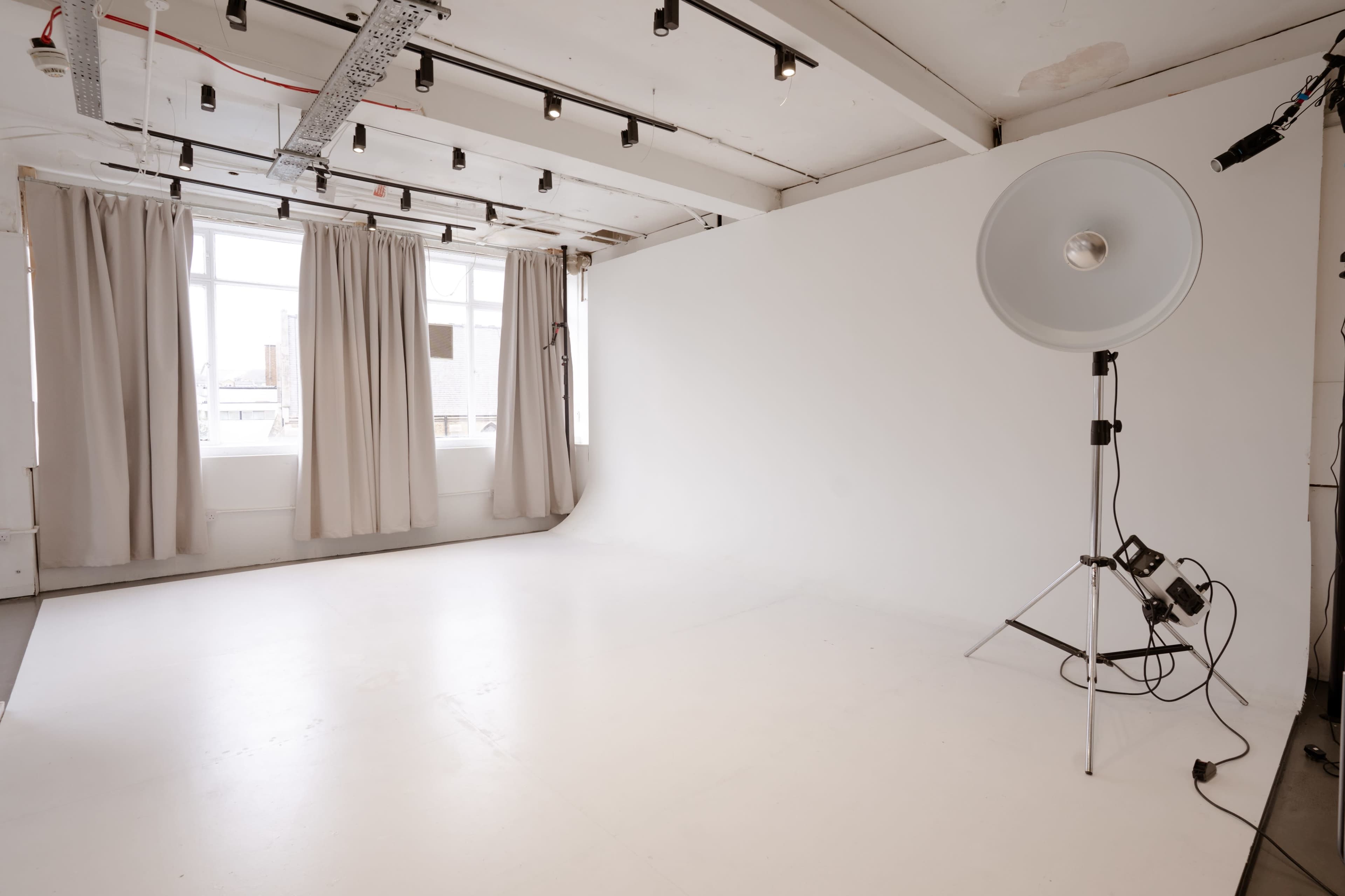 The image shows an empty photography studio with a large light umbrella on a stand next to a white backdrop and partially draped curtains.