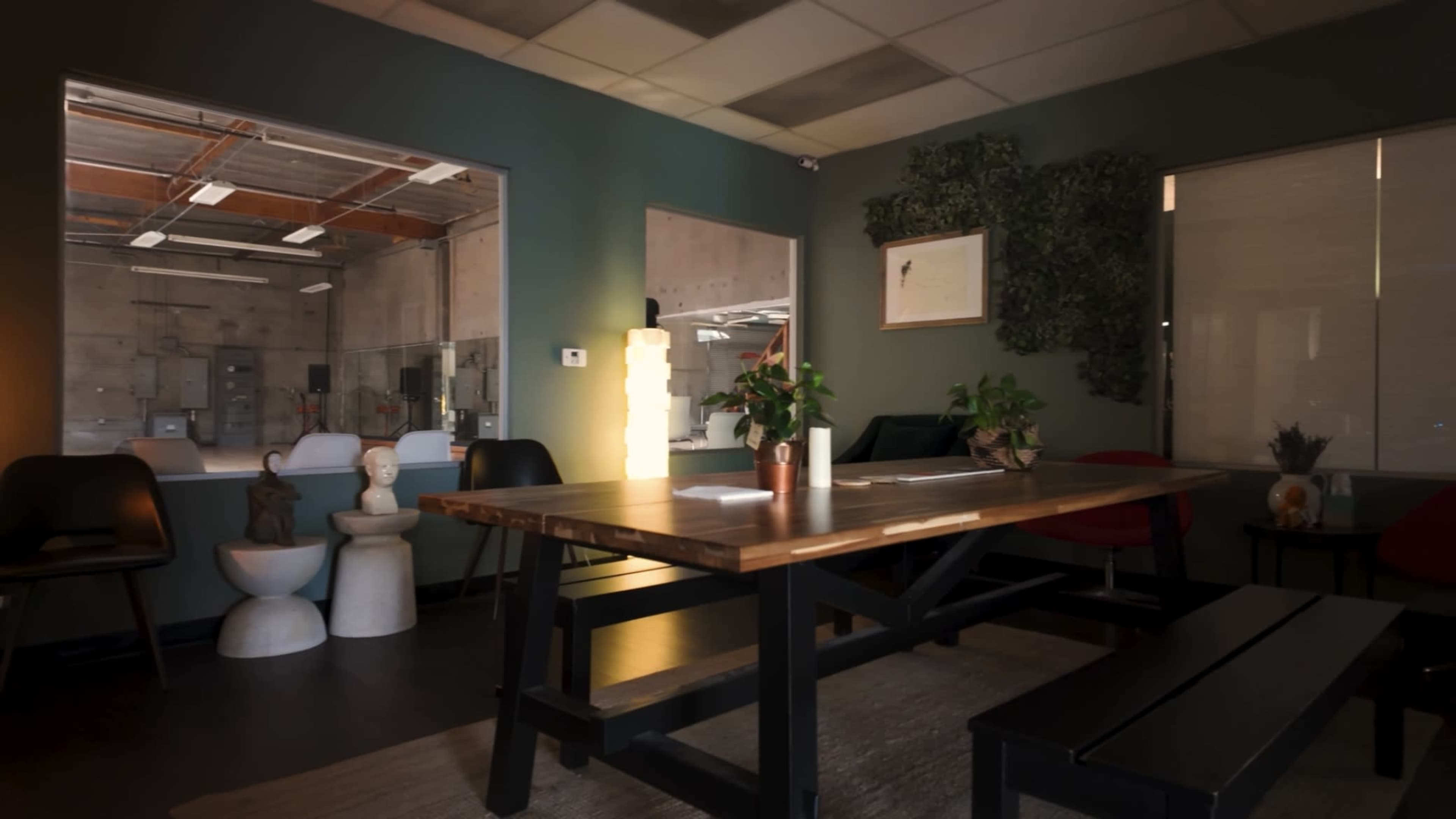 A modern workspace with a wooden table, black chairs, decorative plants, and a large window revealing a concrete interior.